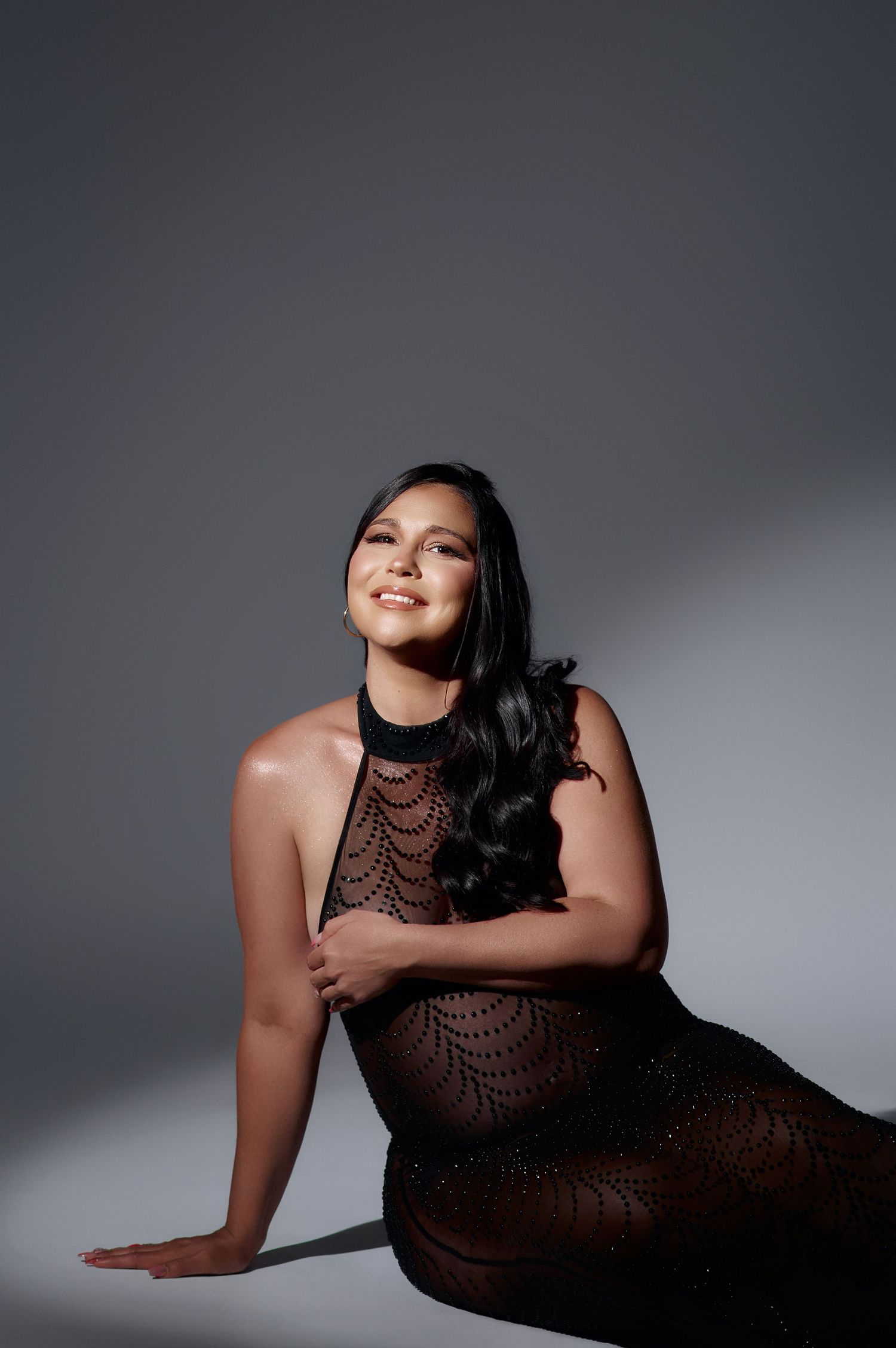 Woman in black lace halter dress sitting gracefully on floor, smiling, with long dark wavy hair against gray background.