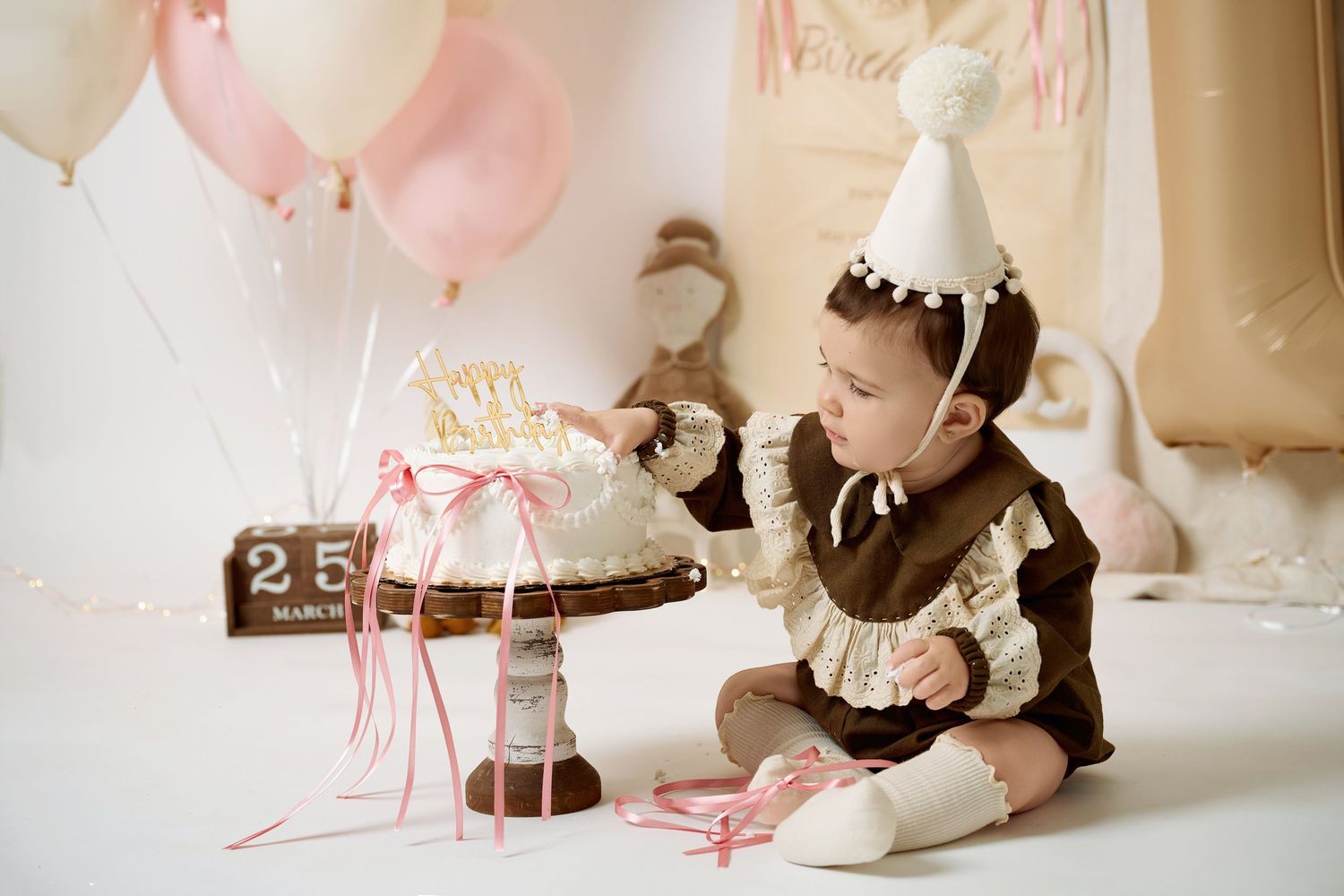 Baby in party hat smashes first birthday cake at a pink and white balloon-decorated cake smash photo shoot.