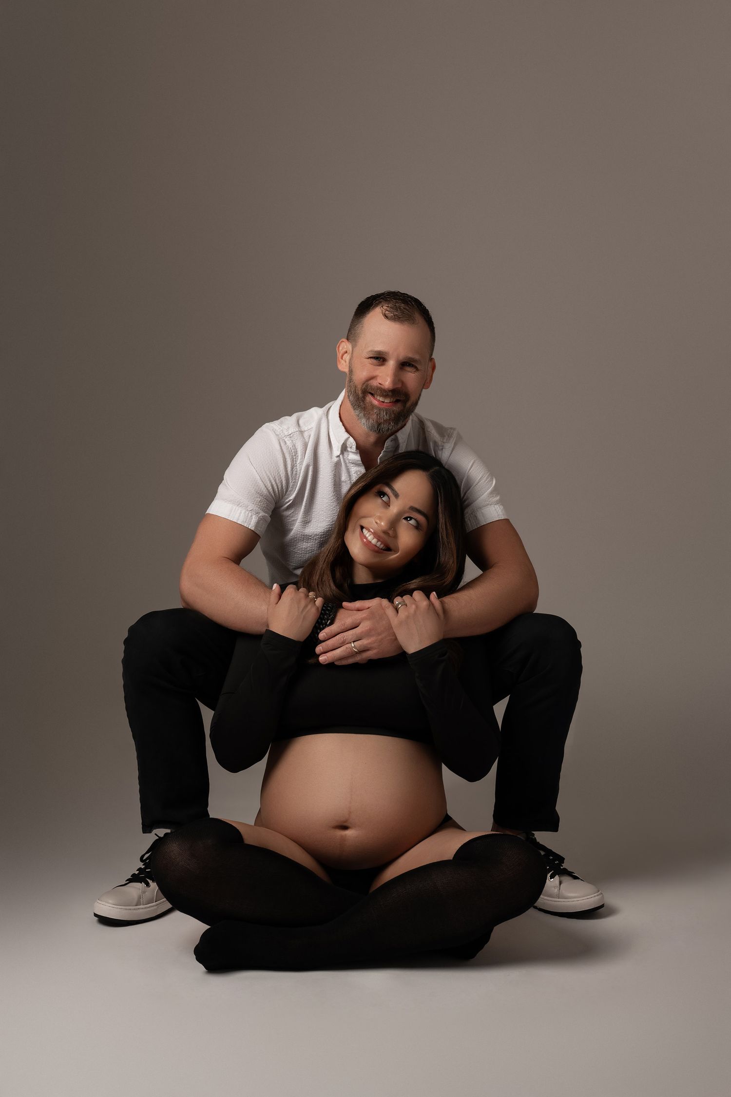 Smiling pregnant couple posing together, man in white shirt embracing woman in black crop top, studio portrait.