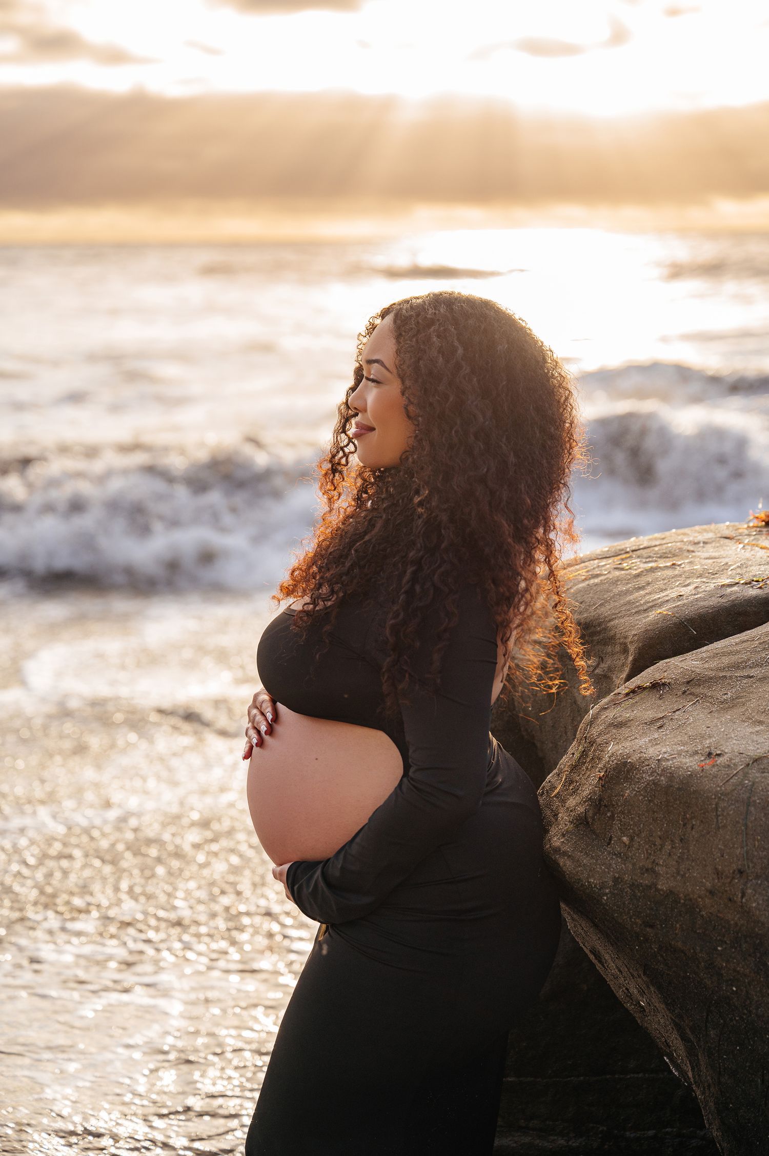 Pregnant woman with curly hair in black dress cradles bump beside coastal rocks during golden sunset.