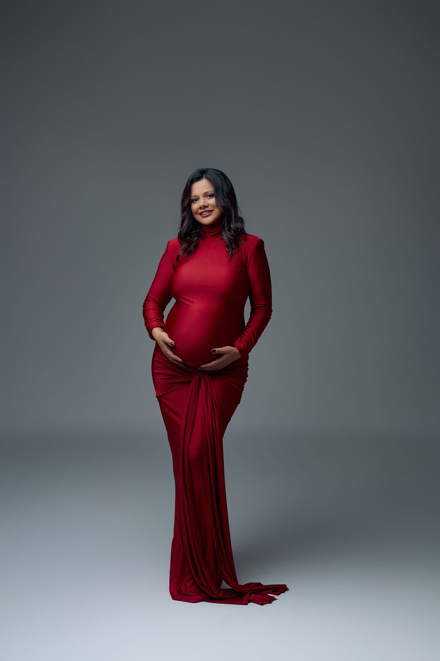 Pregnant woman in elegant red long-sleeve gown cradling baby bump, smiling in professional maternity photo studio shoot.