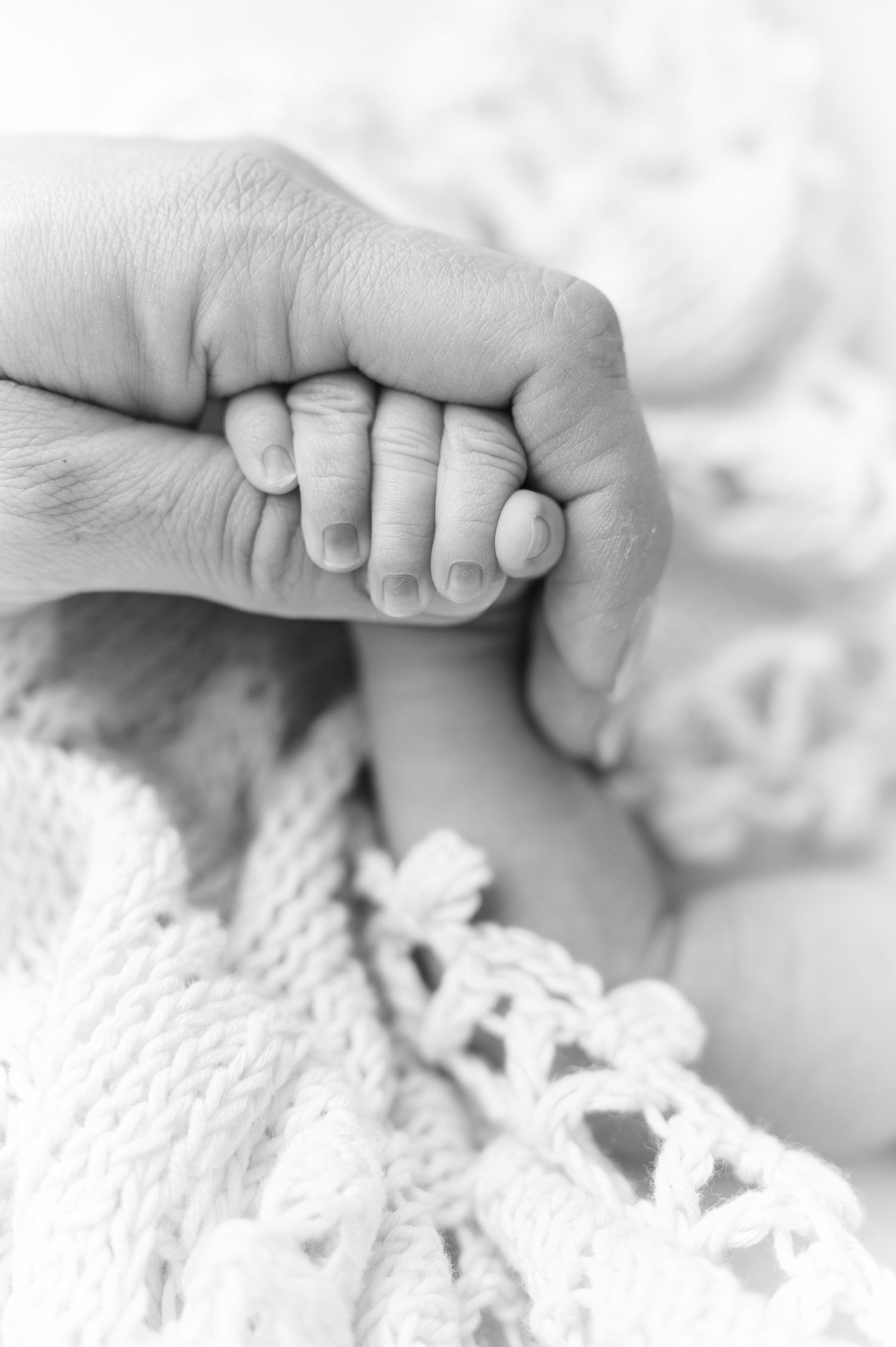 Black and white close-up of a newborn baby grasping an adult finger, lying on a soft knit blanket.