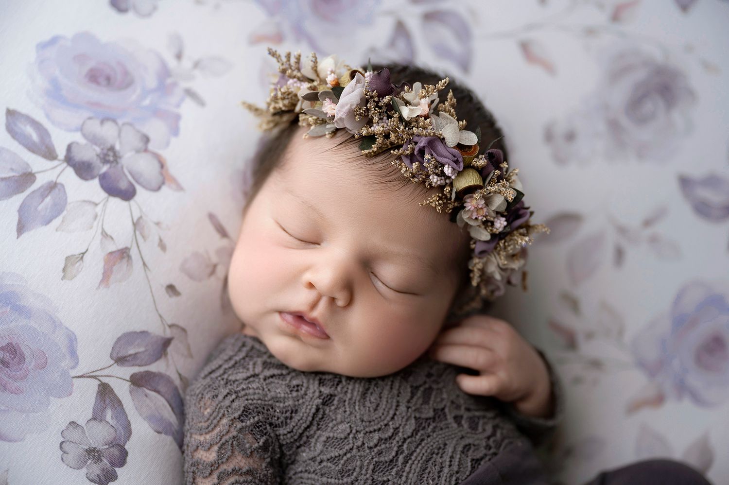 Sleeping newborn baby wearing a delicate floral crown on a soft purple floral backdrop.