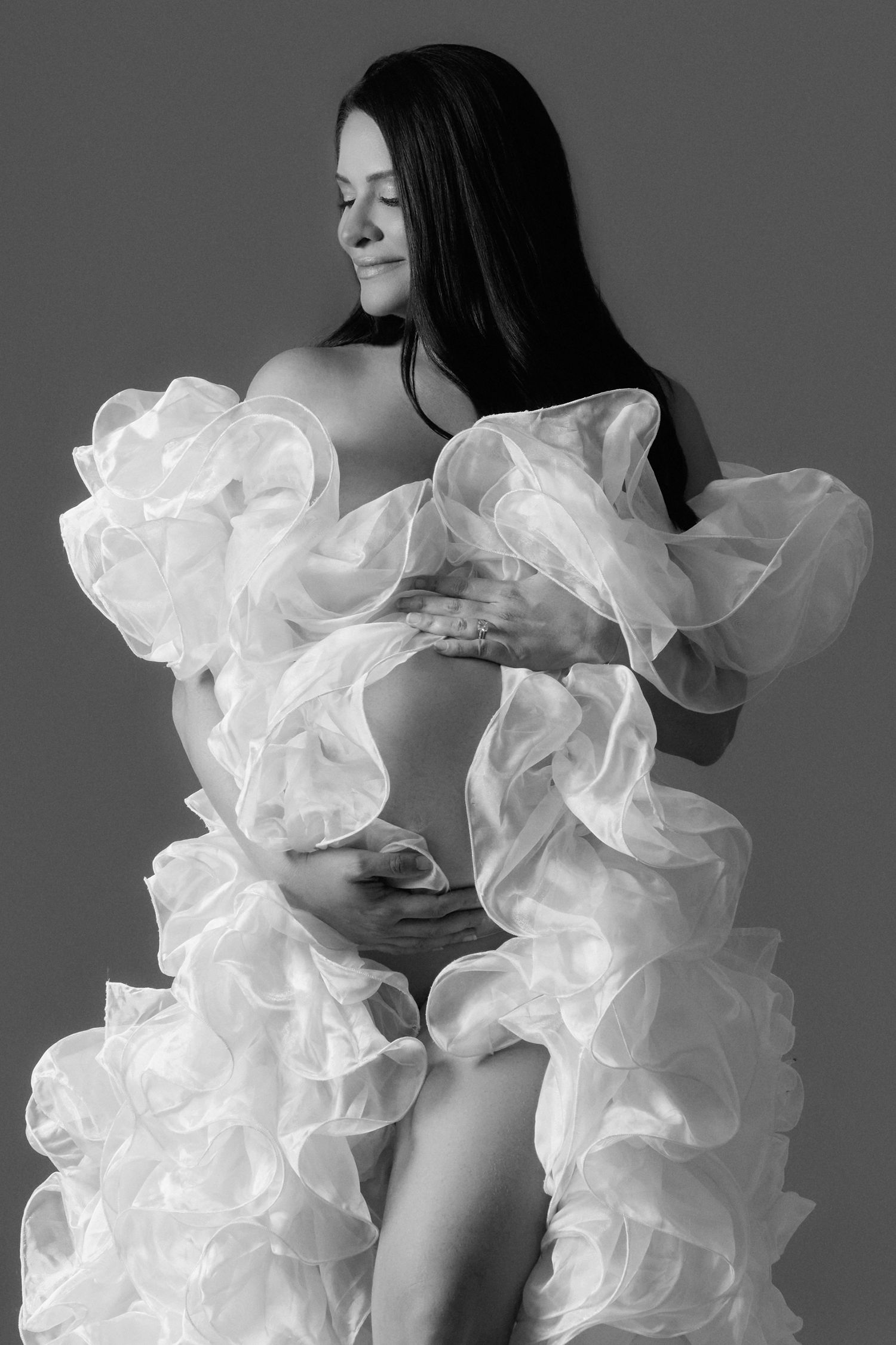 Black and white maternity photo of a pregnant woman in a ruffled white organza gown, cradling her baby bump.