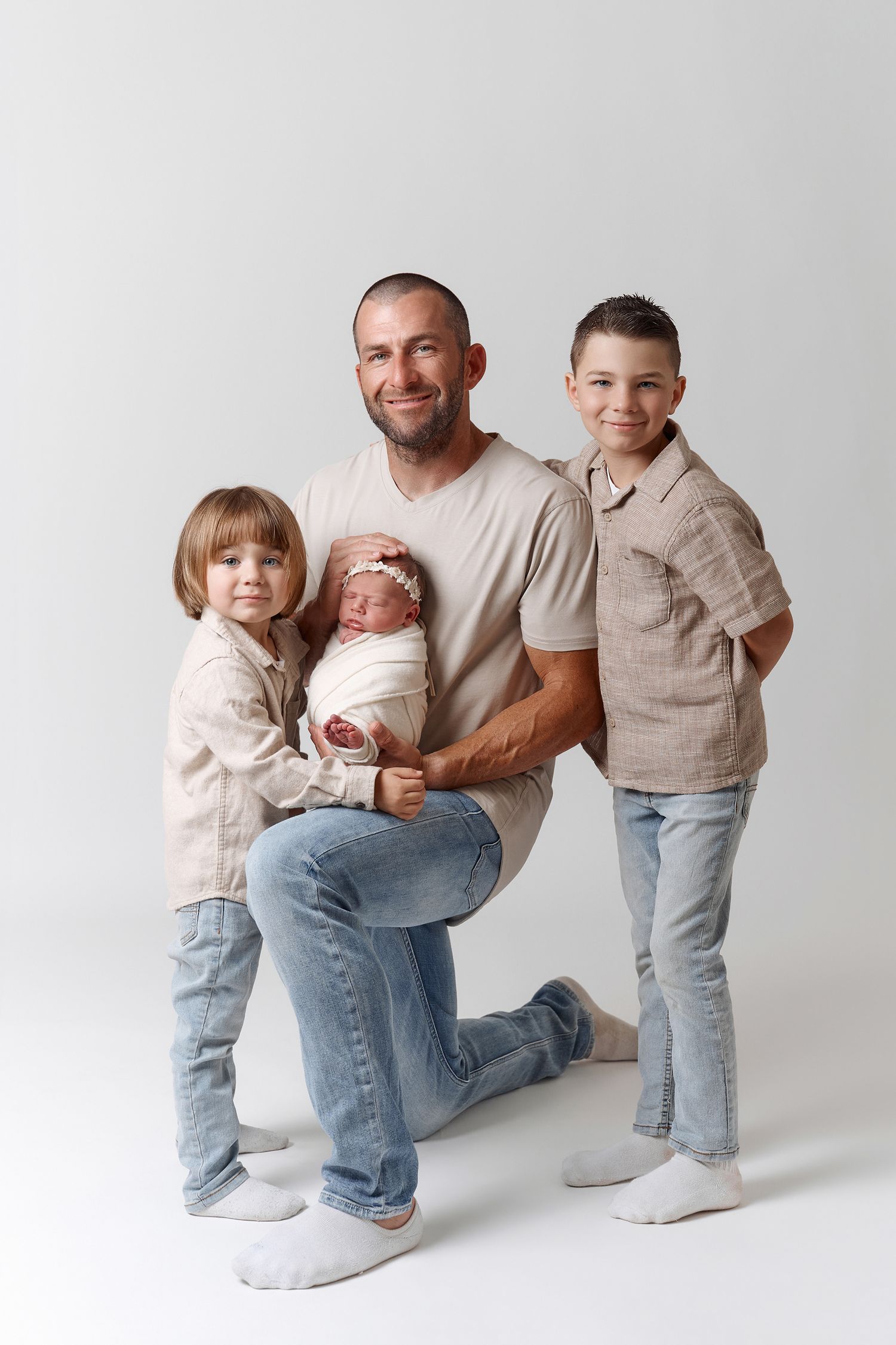 Father kneeling with three children, holding newborn baby, flanked by toddler girl and older boy, studio photo.