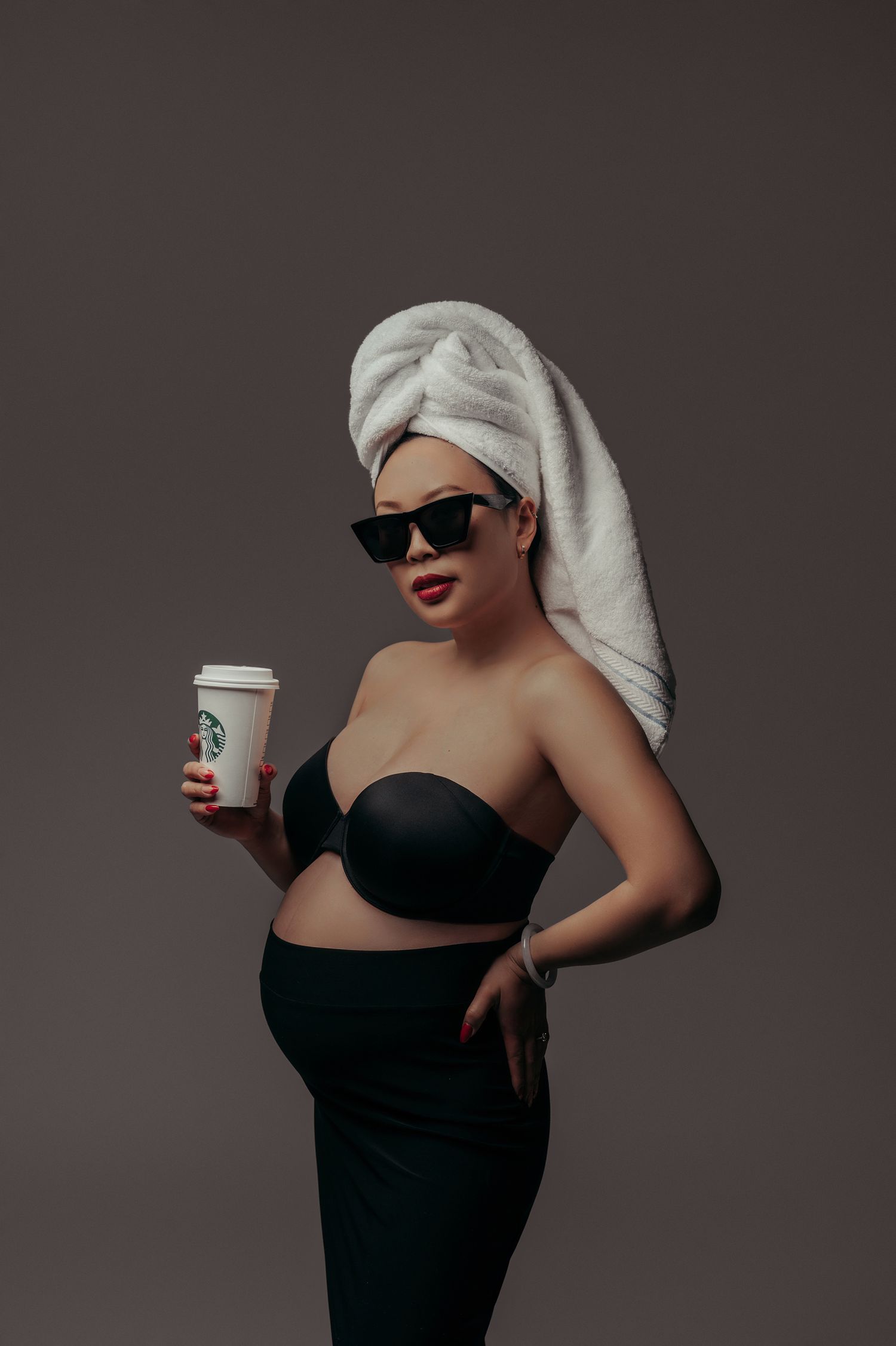Pregnant woman in black strapless top, sunglasses, and towel turban holding a Starbucks cup on gray background.