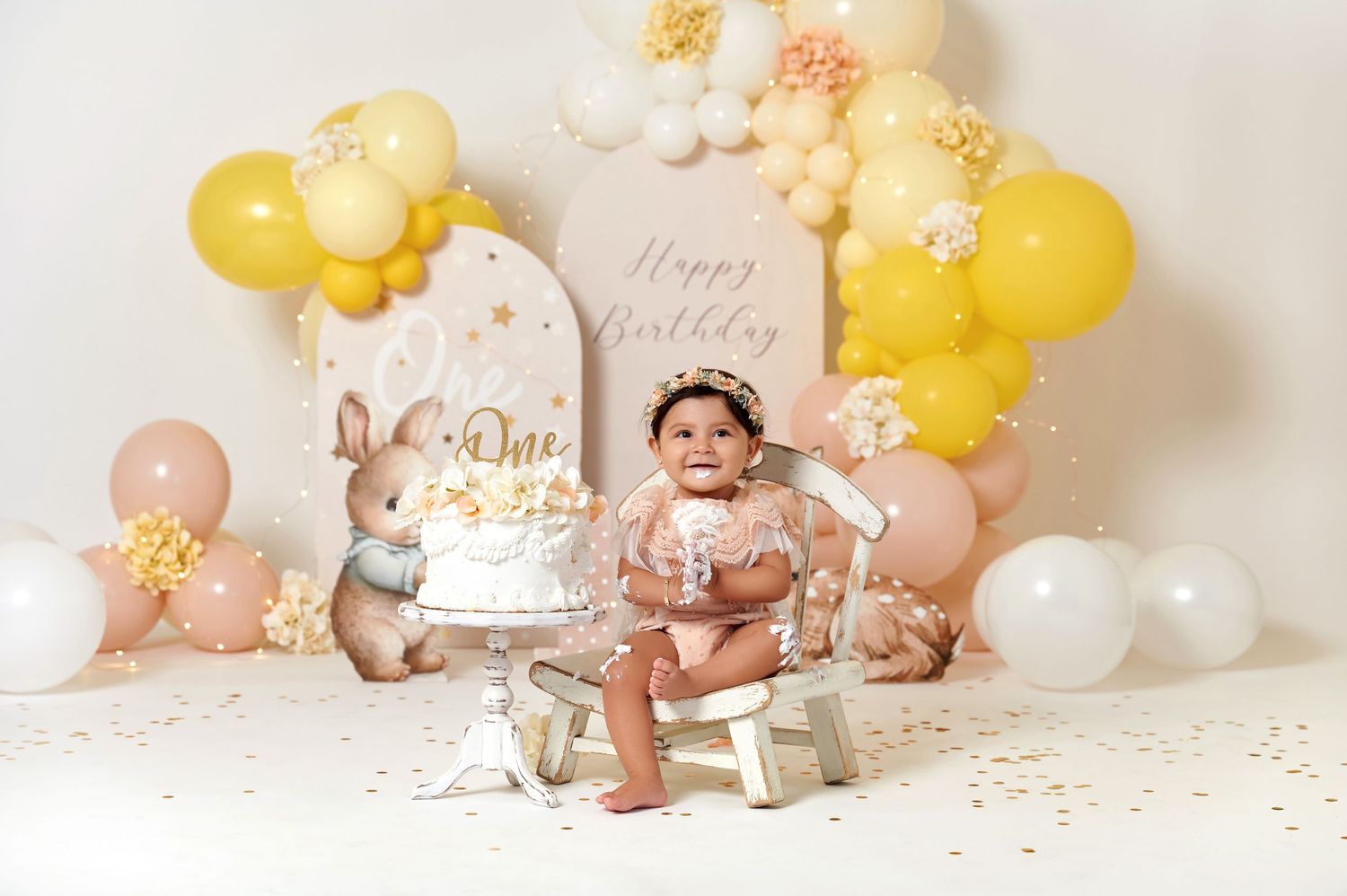 Baby's first birthday cake smash with bunny decorations, yellow and pink balloon garland, and Happy Birthday backdrop.