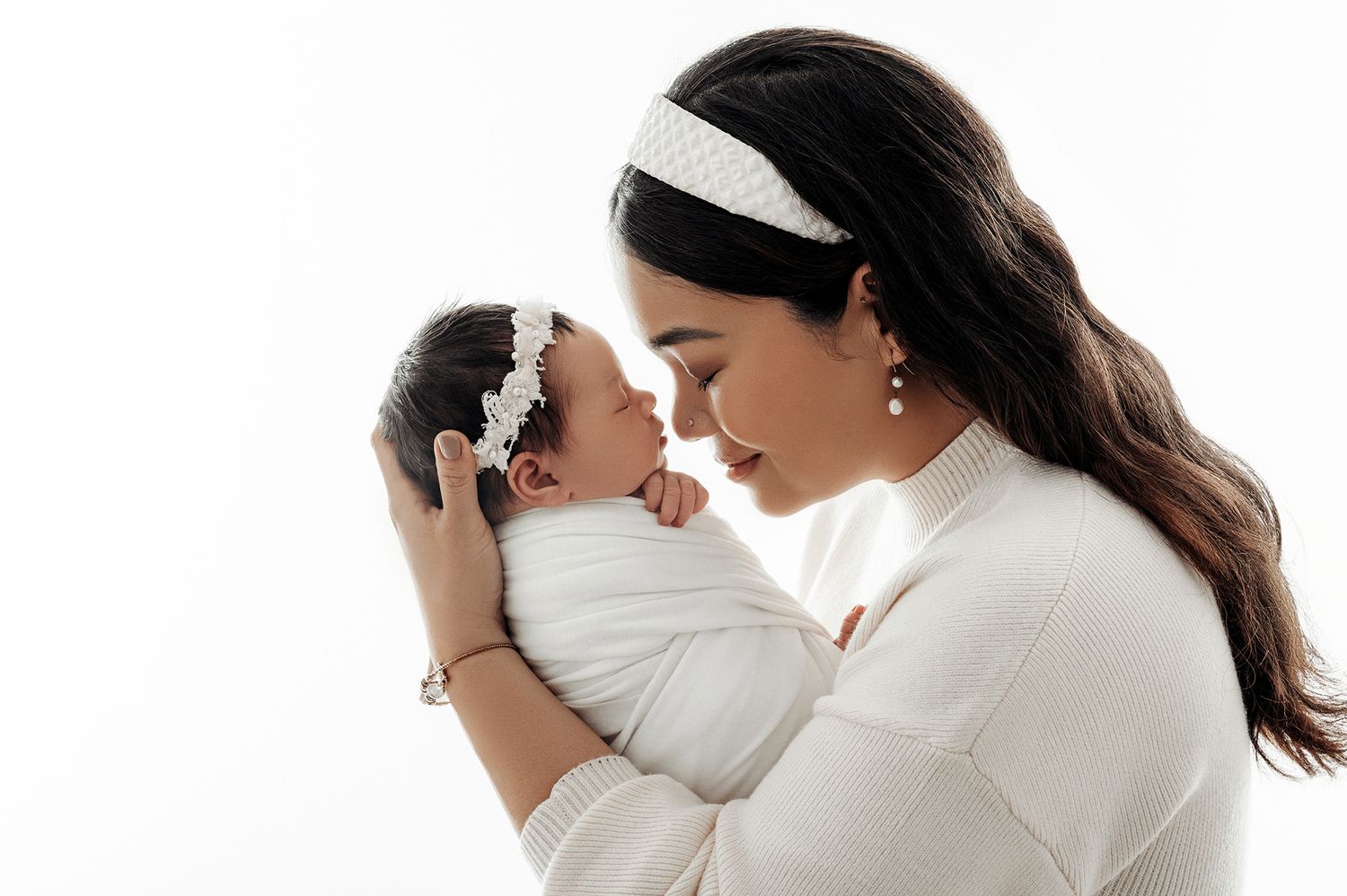 Mother in white sweater tenderly holds swaddled newborn baby, both wearing delicate headbands, against white background.