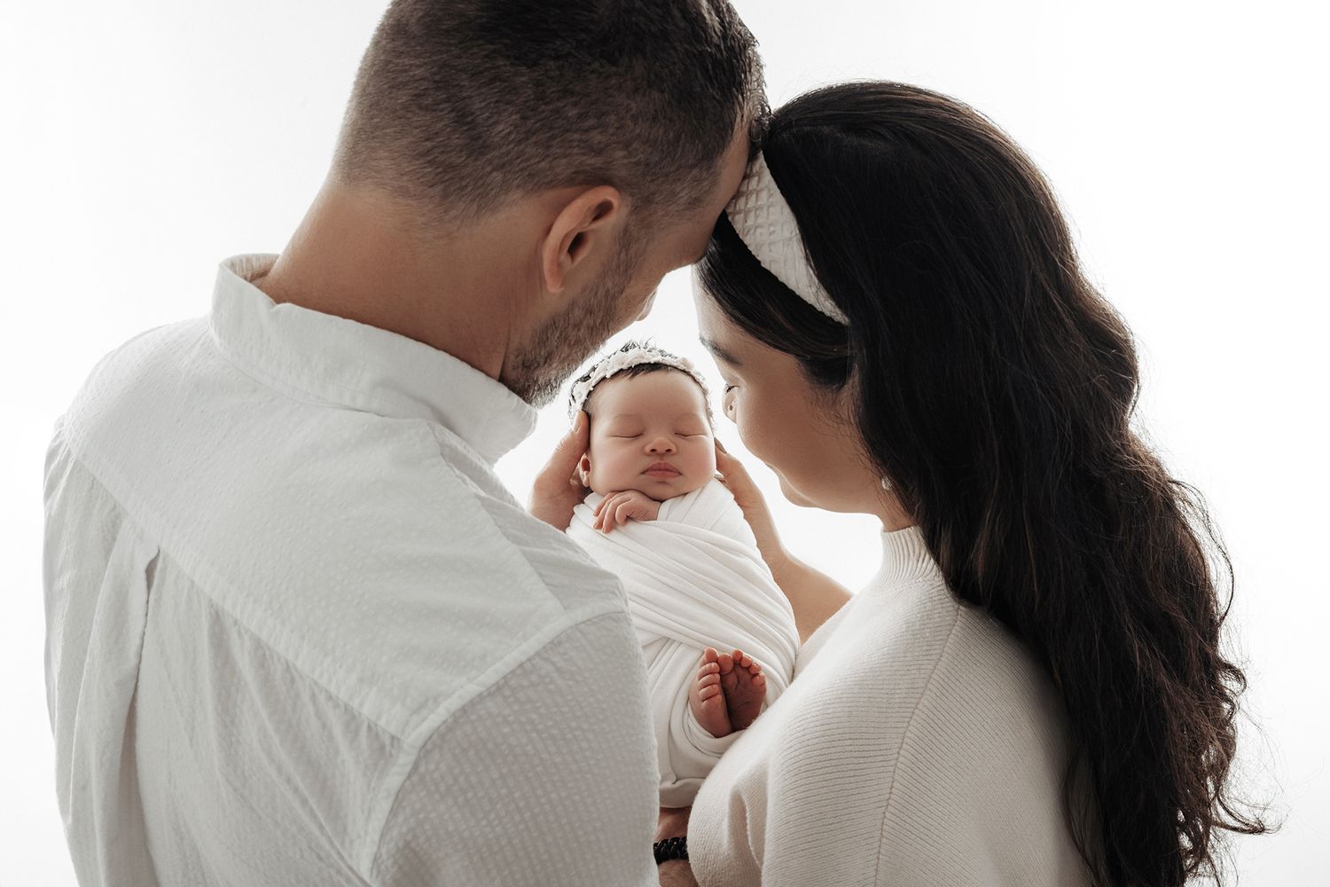Parents in white clothing tenderly hold their sleeping newborn baby wrapped in white against a bright background.