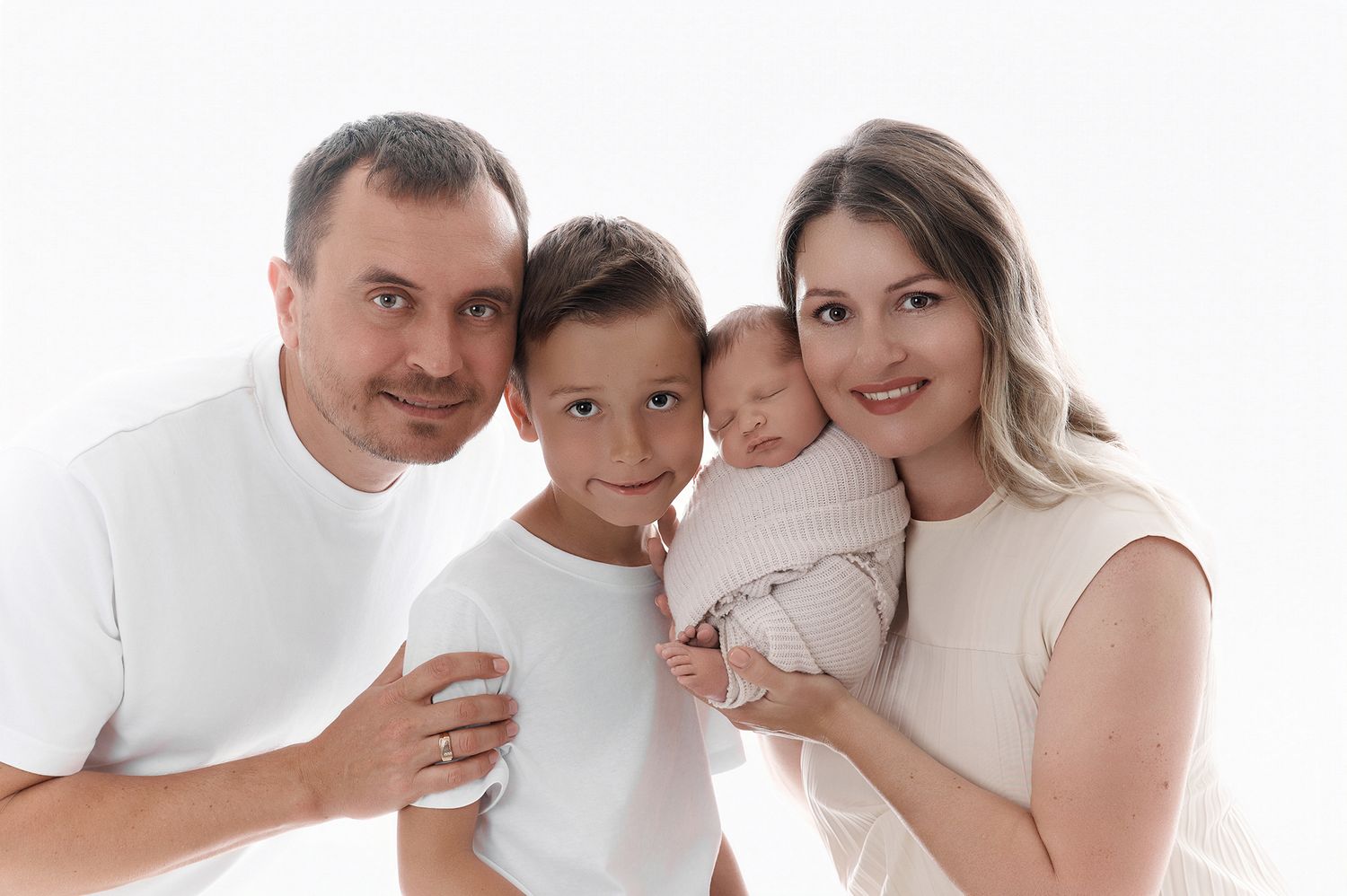 Happy family of four posing together, parents with young son and newborn baby, wearing white outfits on white background.