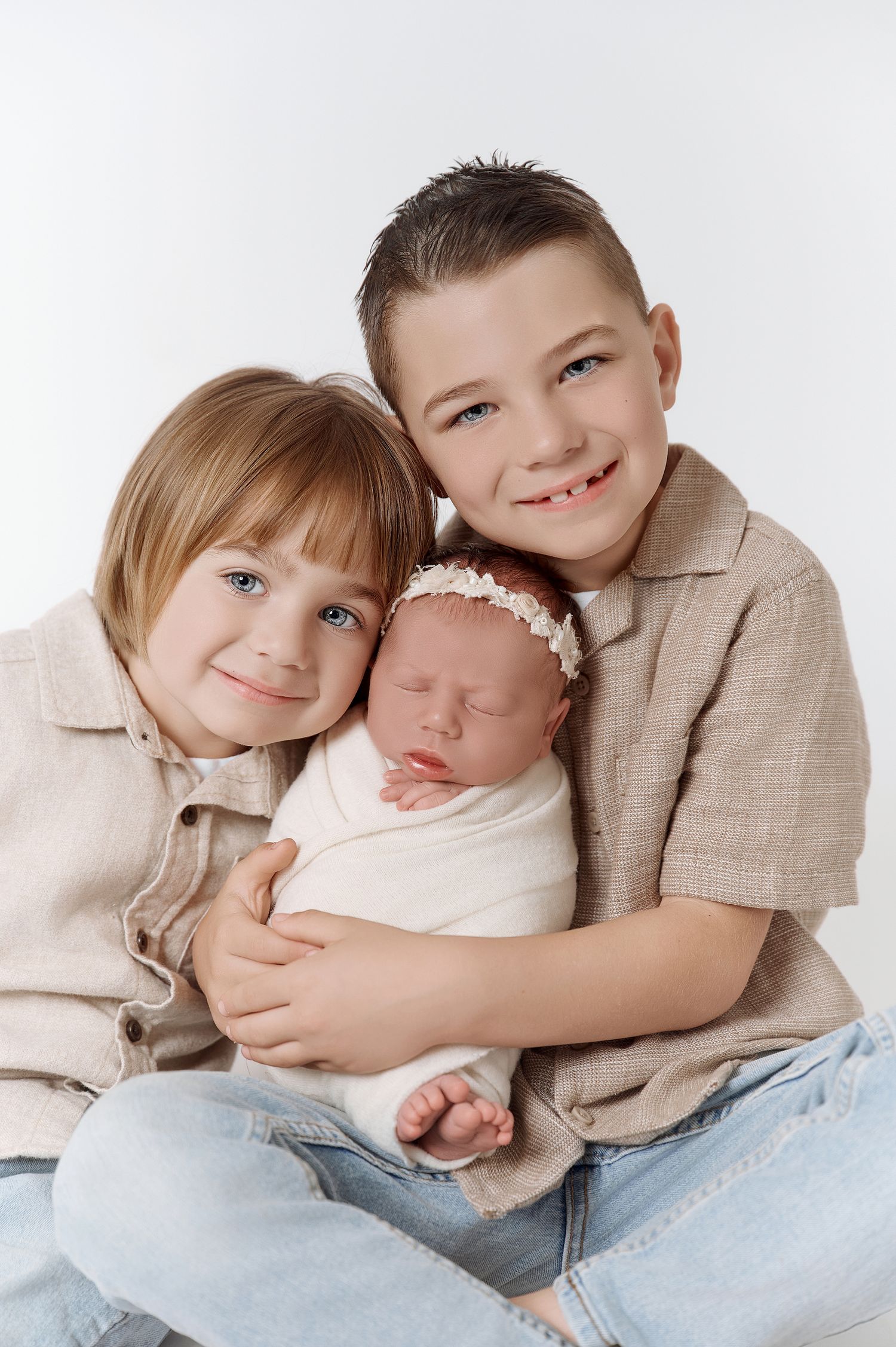 Two older siblings lovingly hold their sleeping newborn baby sister in a bright white studio portrait.