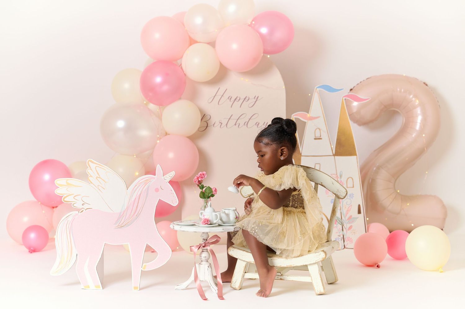 Toddler girl in gold dress at unicorn-themed 2nd birthday setup with pink balloons and Happy Birthday backdrop.