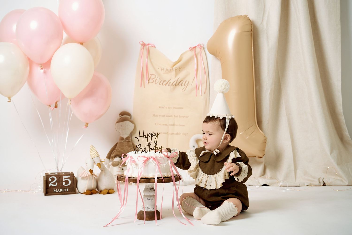 Baby girl celebrates first birthday with pink balloons, cake, and a gold number one balloon in a soft studio setting.