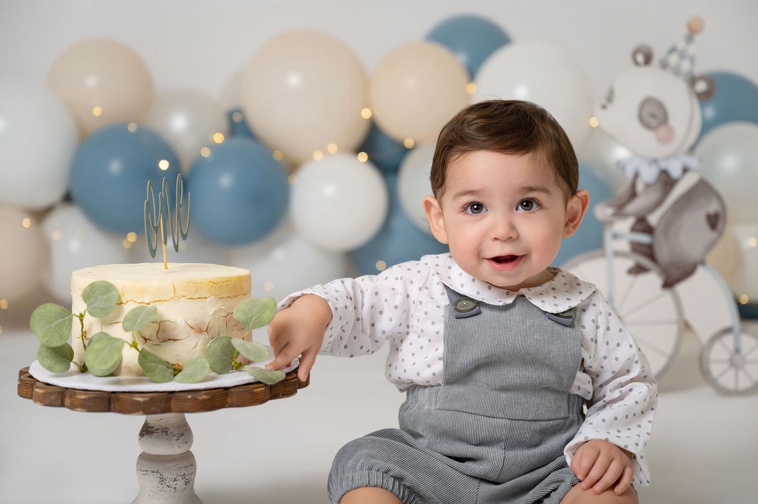 Baby boy in gray overalls reaching for a first birthday cake surrounded by blue and white balloons.
