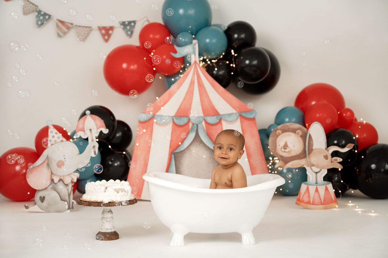 Baby sits in white bathtub at circus-themed first birthday cake smash with red, black, and teal balloon decorations.