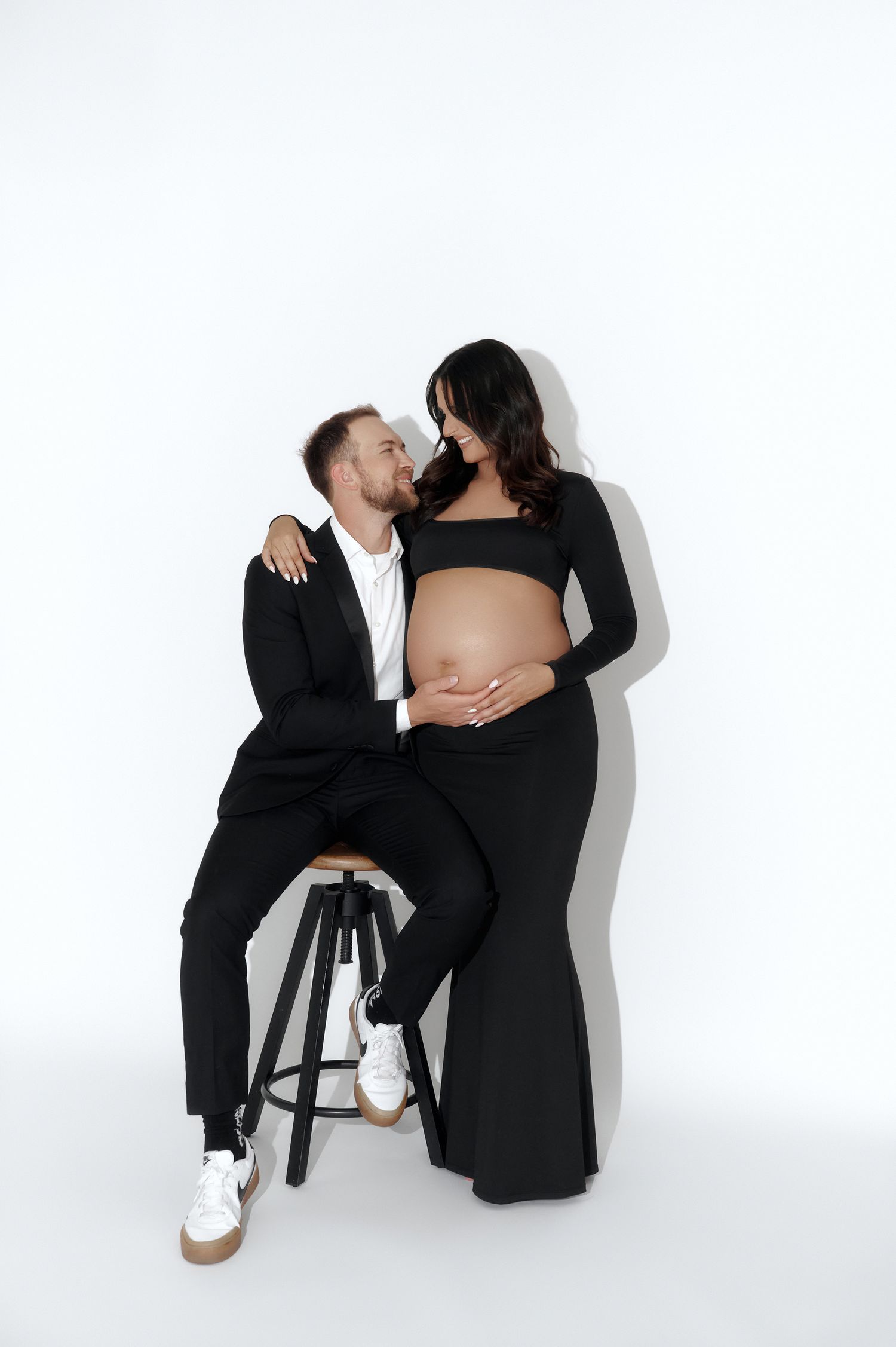 Couple in black outfits posing together, pregnant woman standing while man sits on stool kissing her cheek.