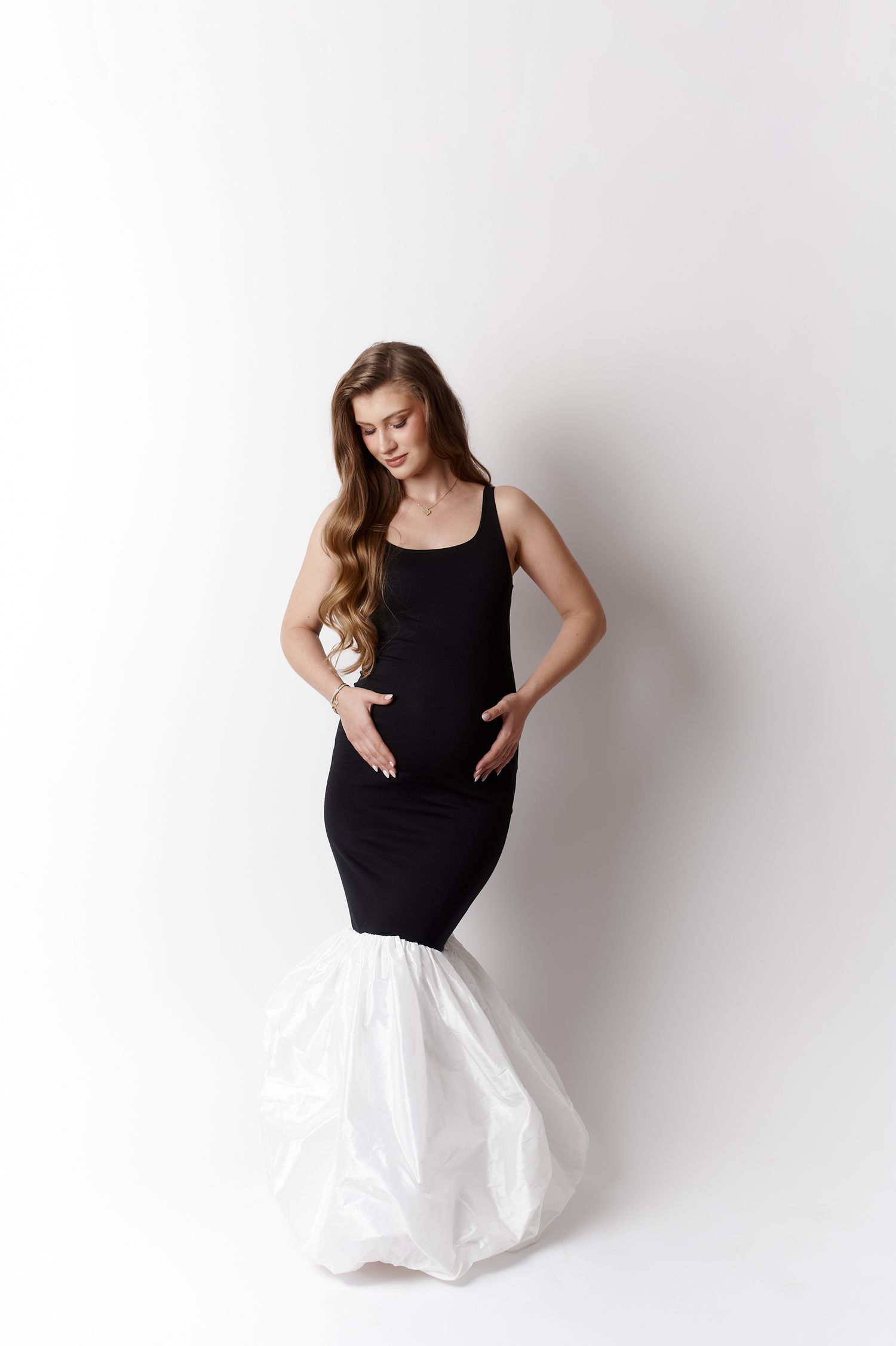 Pregnant woman in black fitted dress with white ruffled hem poses gracefully against white background.