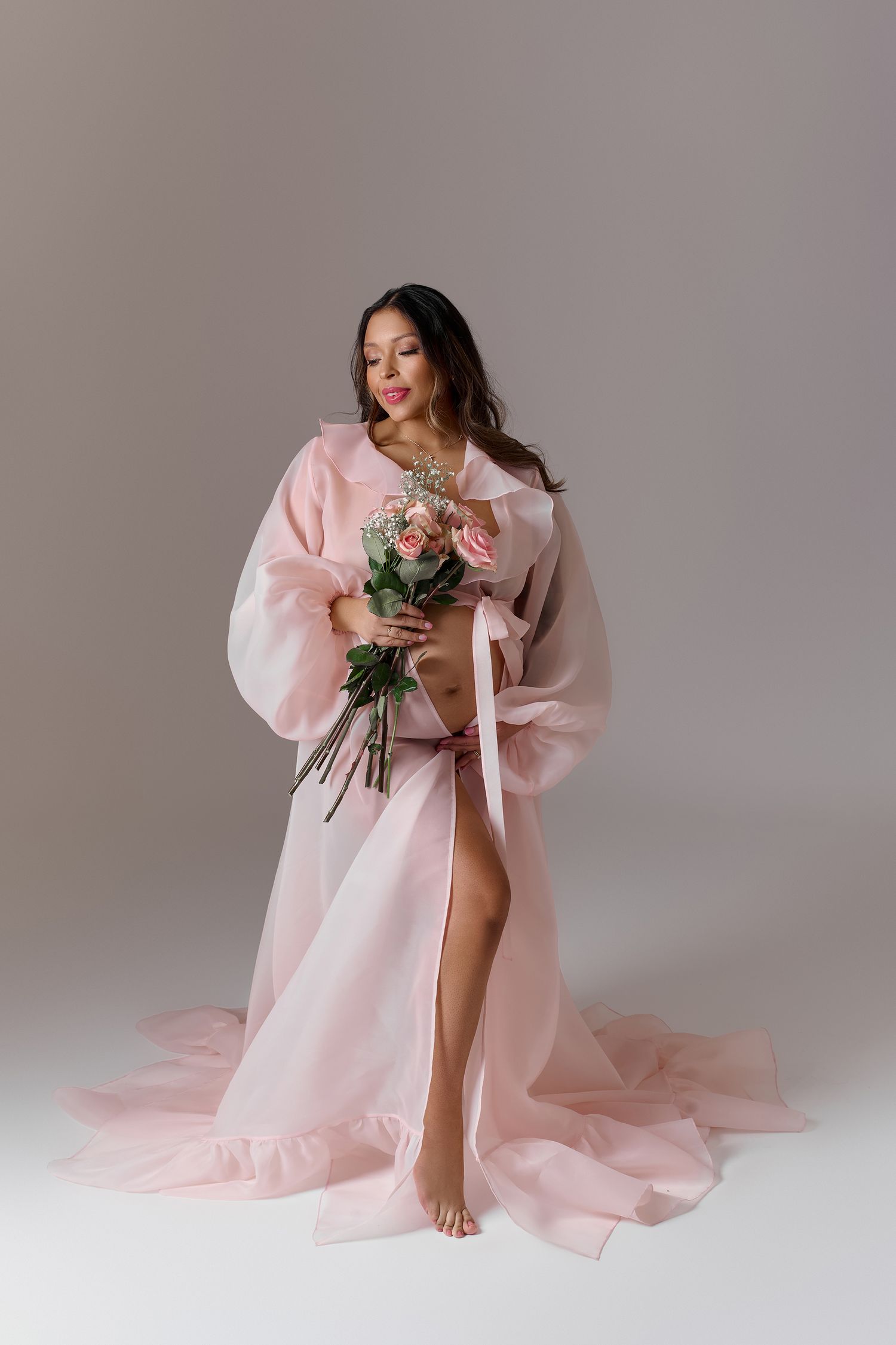 Woman in flowing pink sheer robe holding a bouquet of pink flowers, posing gracefully on a light gray background.