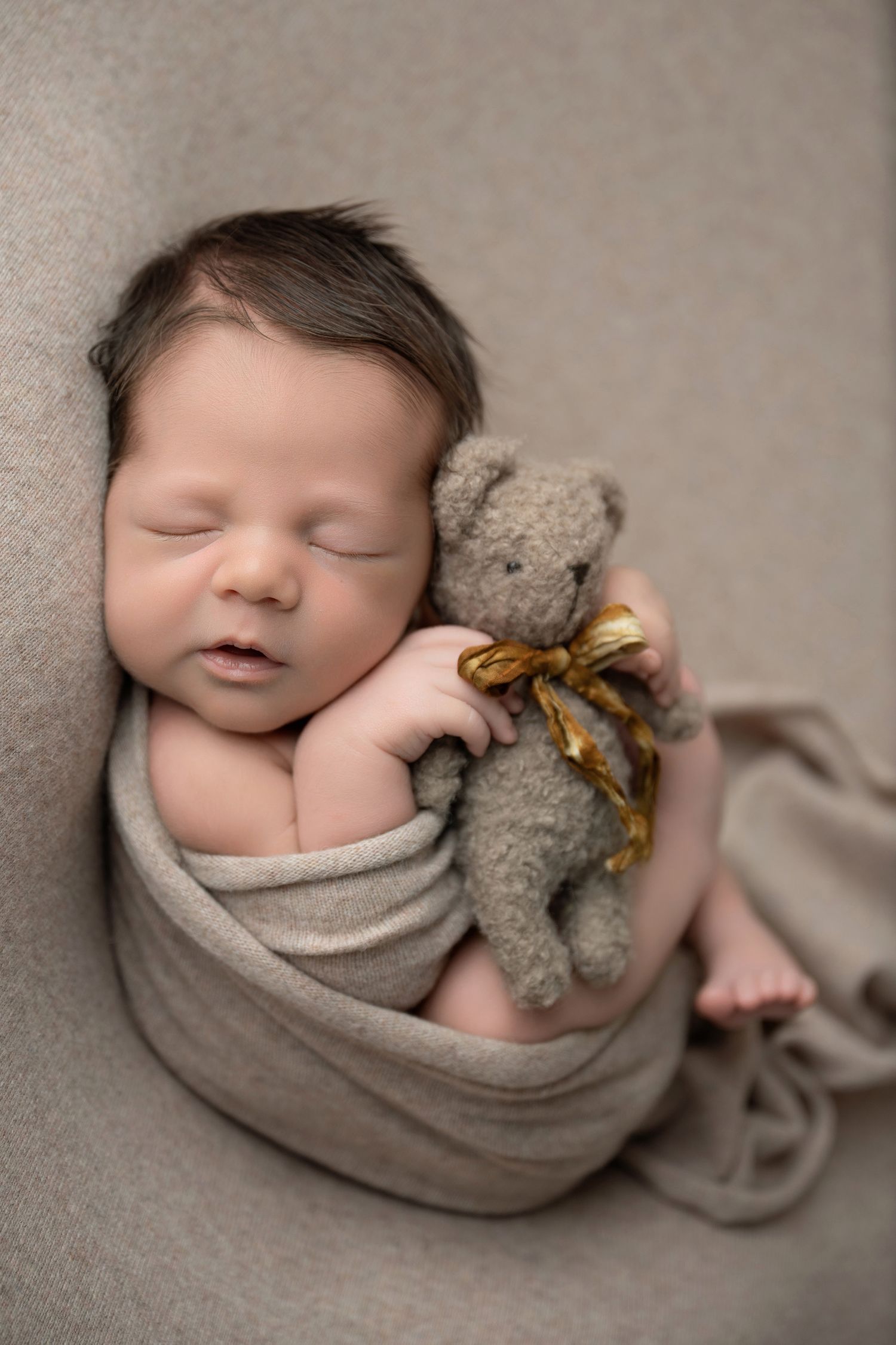 Sleeping newborn baby swaddled in gray wrap holding a tiny teddy bear with gold ribbon bow.