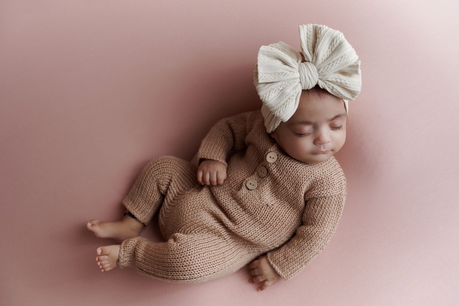 Sleeping newborn baby in a tan knit romper and cream bow headband on a pink background.