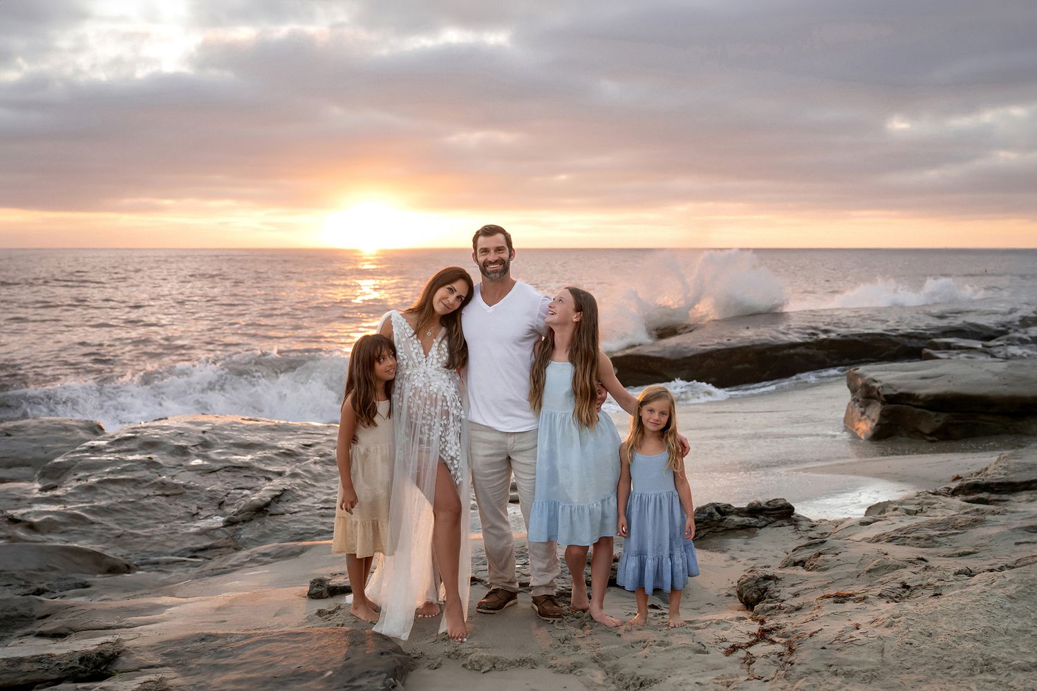 Family of five posing on rocky beach at vibrant sunset with crashing ocean waves behind them.