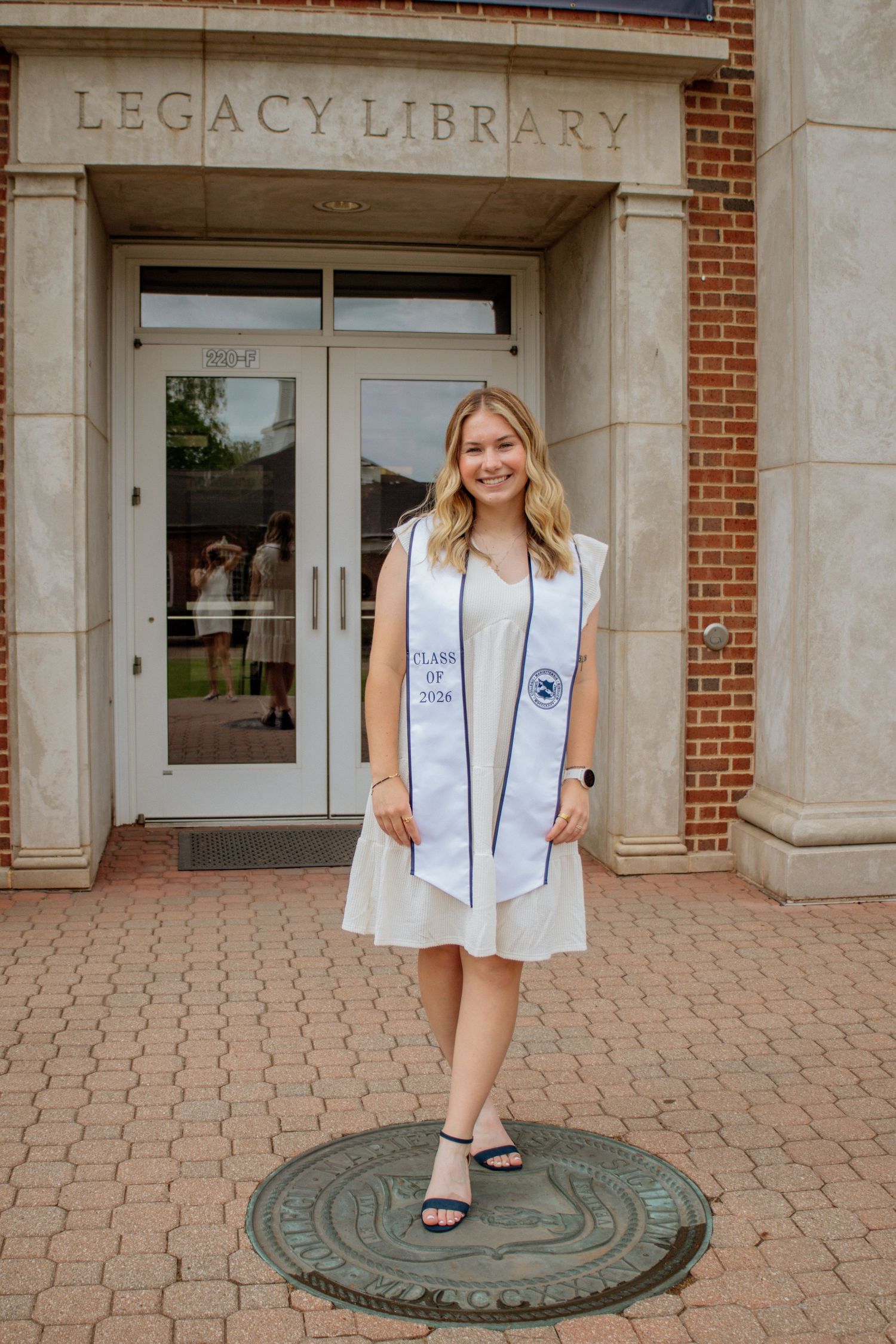 Smiling blonde graduate in white dress with Class of 2026 stole stands on university seal outside Legacy Library.