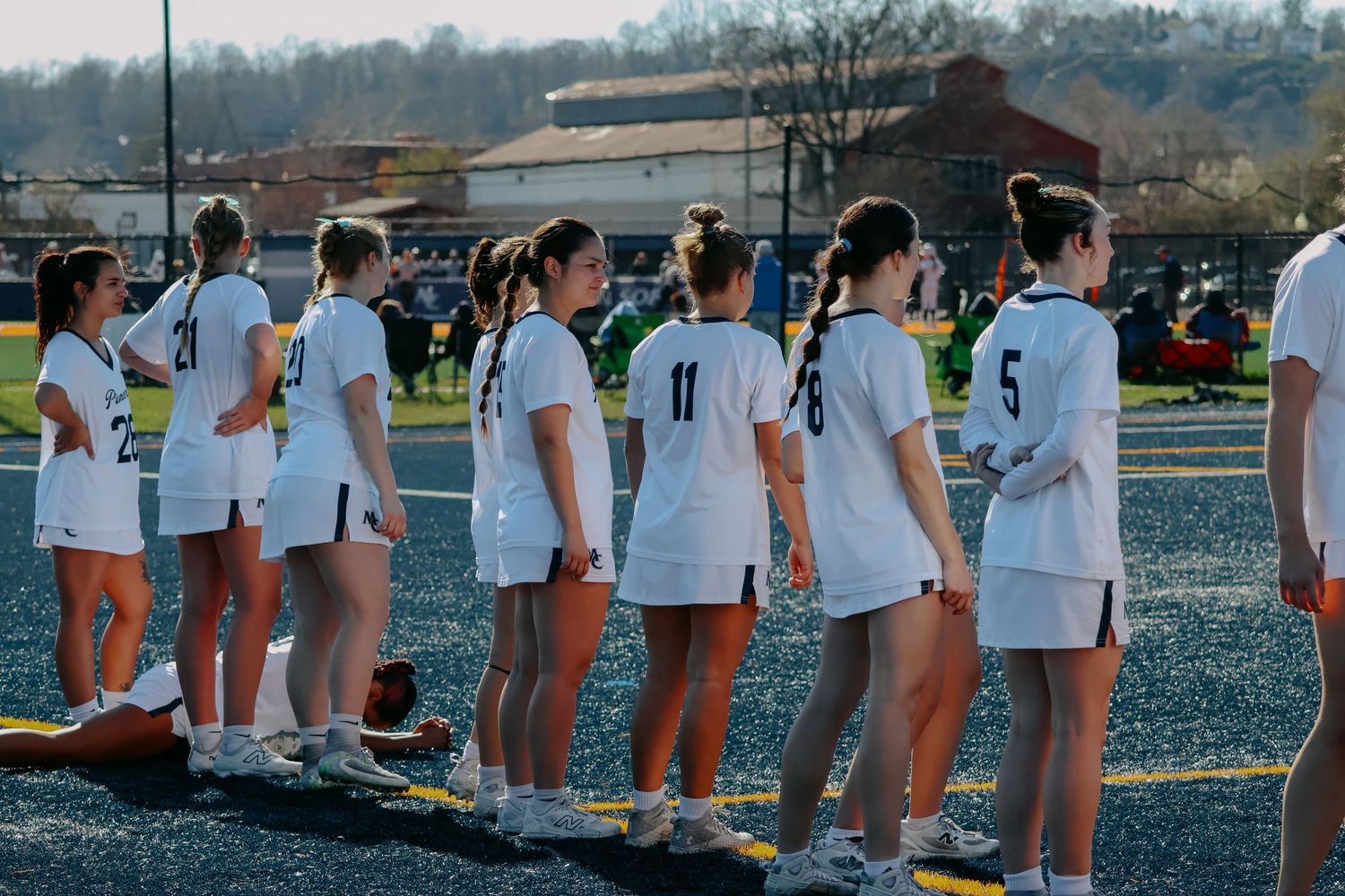 Female lacrosse team in white uniforms lines up on a turf field before a game.