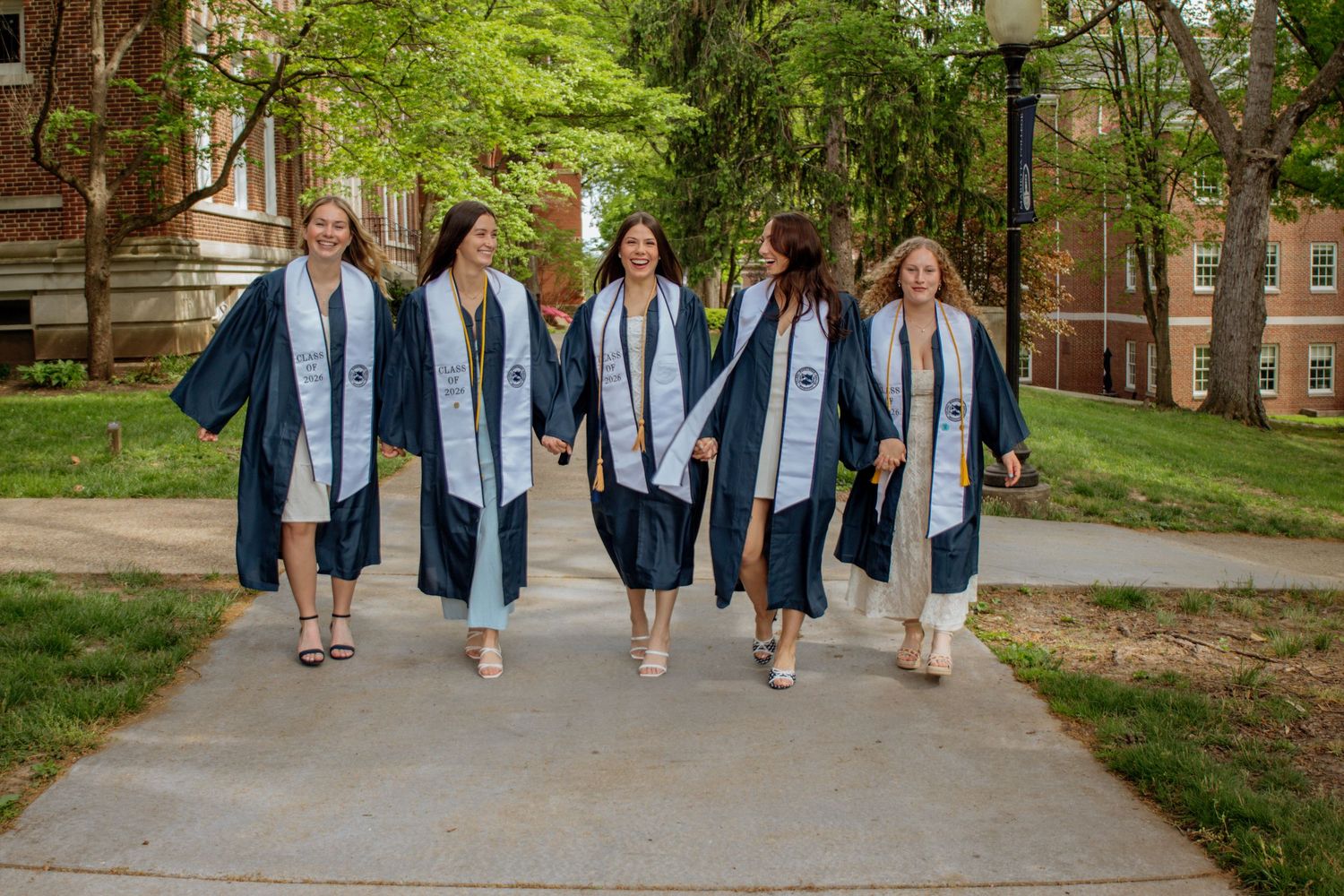 Six female graduates walk together in navy blue graduation gowns along a campus pathway.