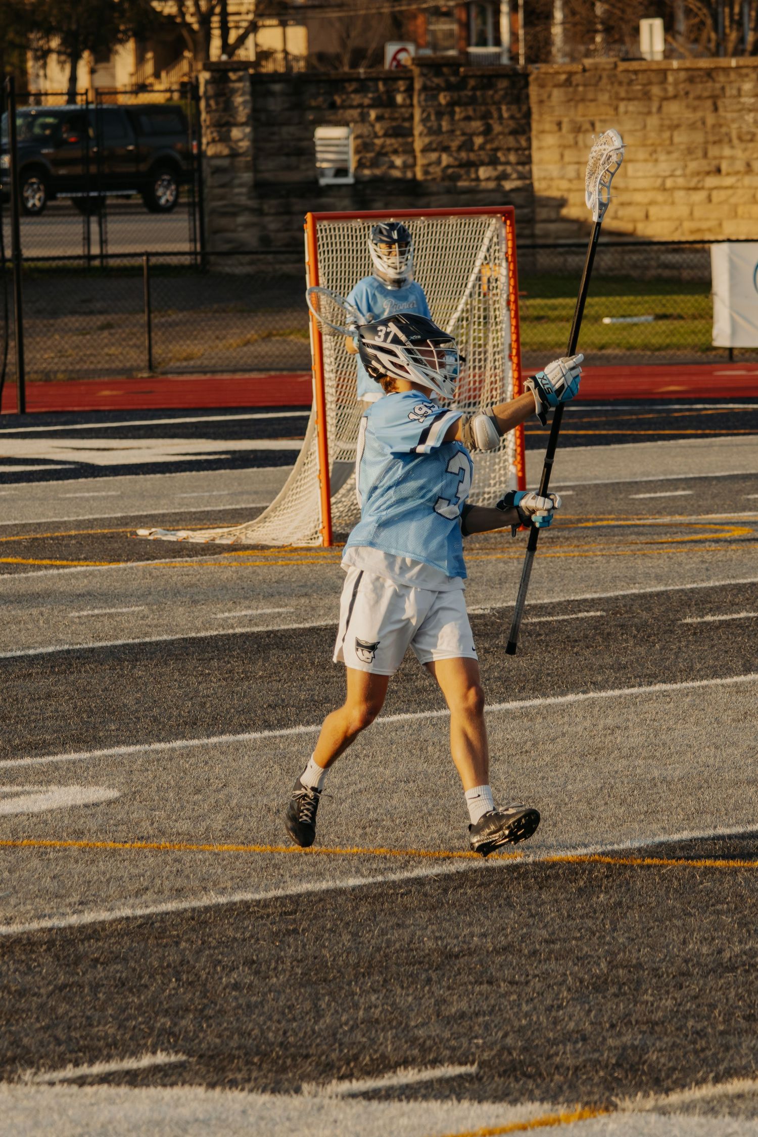 A lacrosse player in a light blue jersey holds a stick near a goal net on an outdoor court during a sunny day.