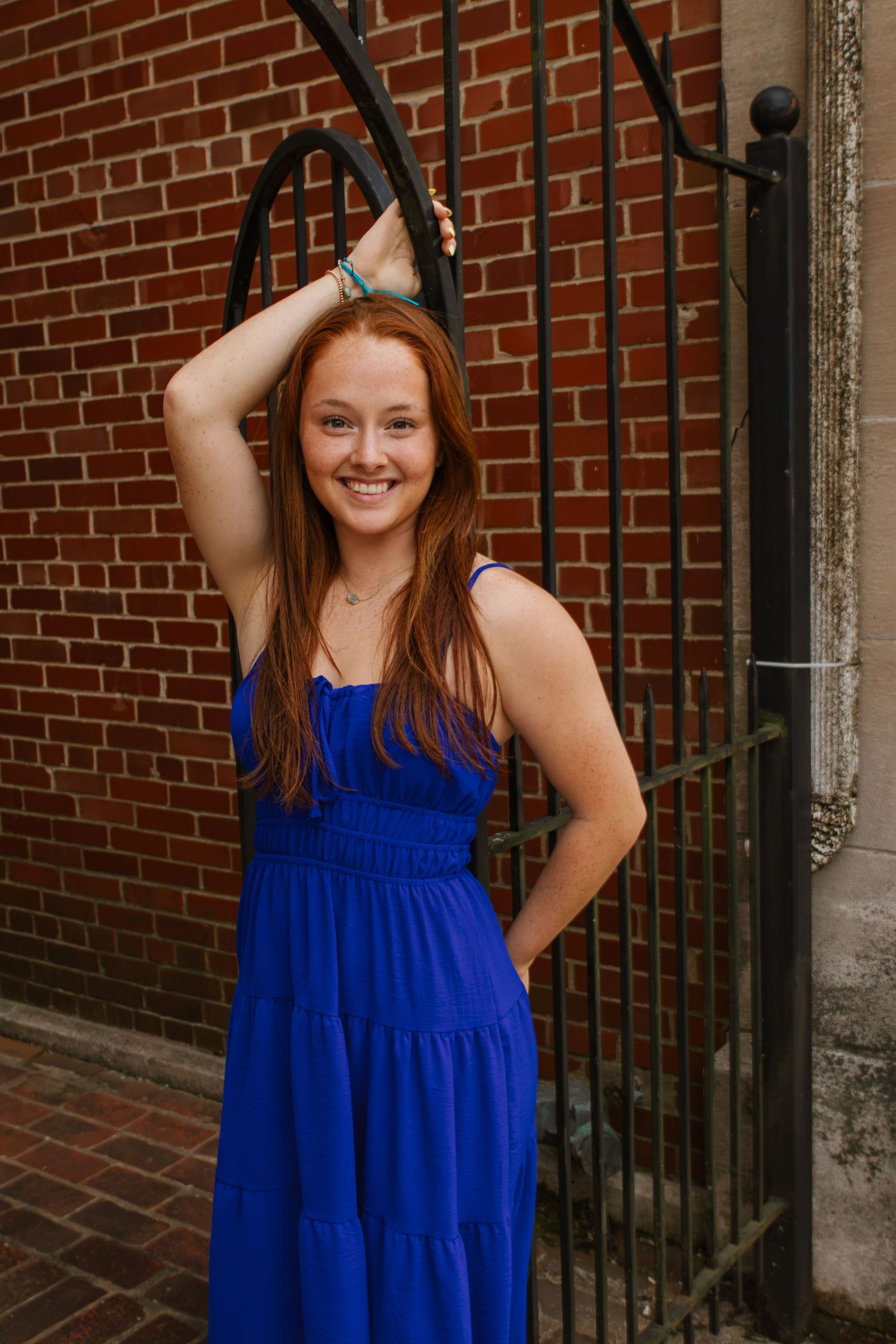 Smiling young woman with red hair in a blue sundress posing by a black iron fence against a brick wall.