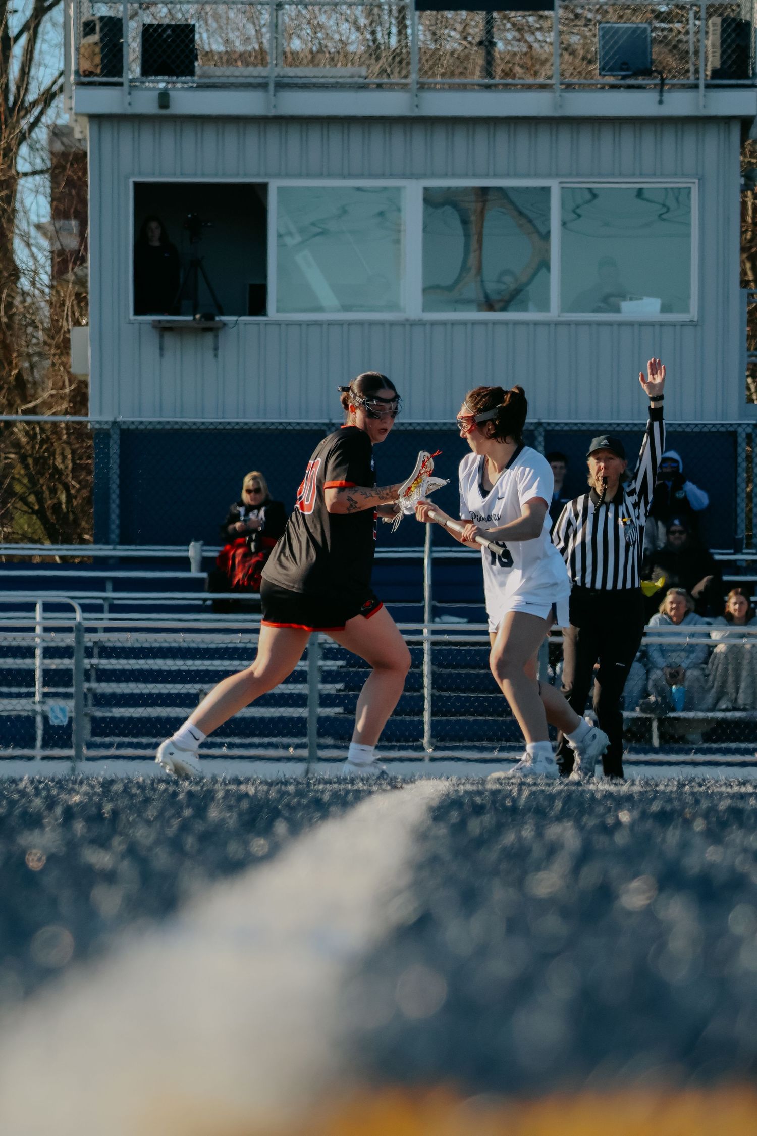 Female lacrosse player in white uniform battles an opponent while a referee signals nearby.