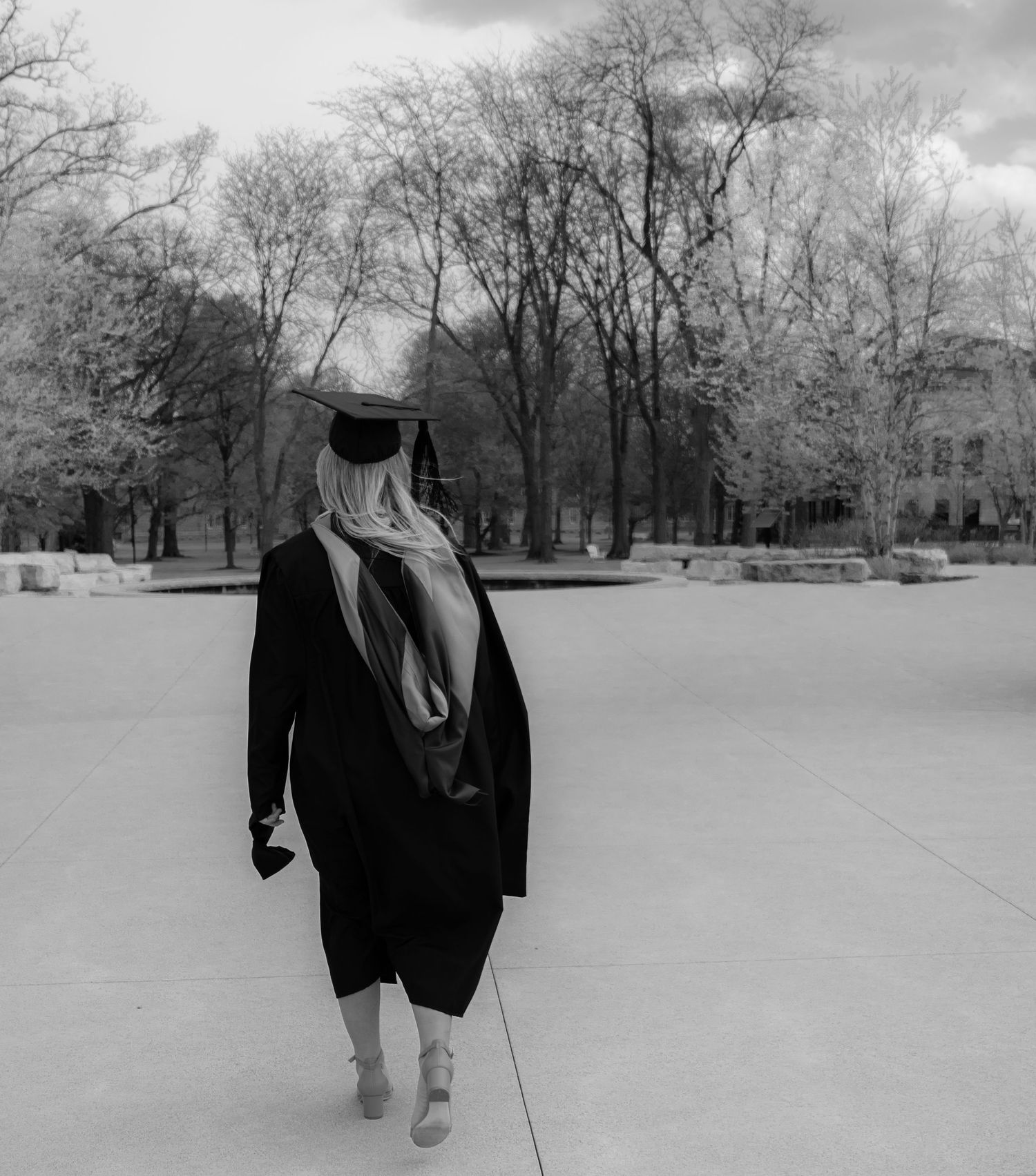 Black and white photo of a graduate in cap and gown walking across a campus plaza alone.