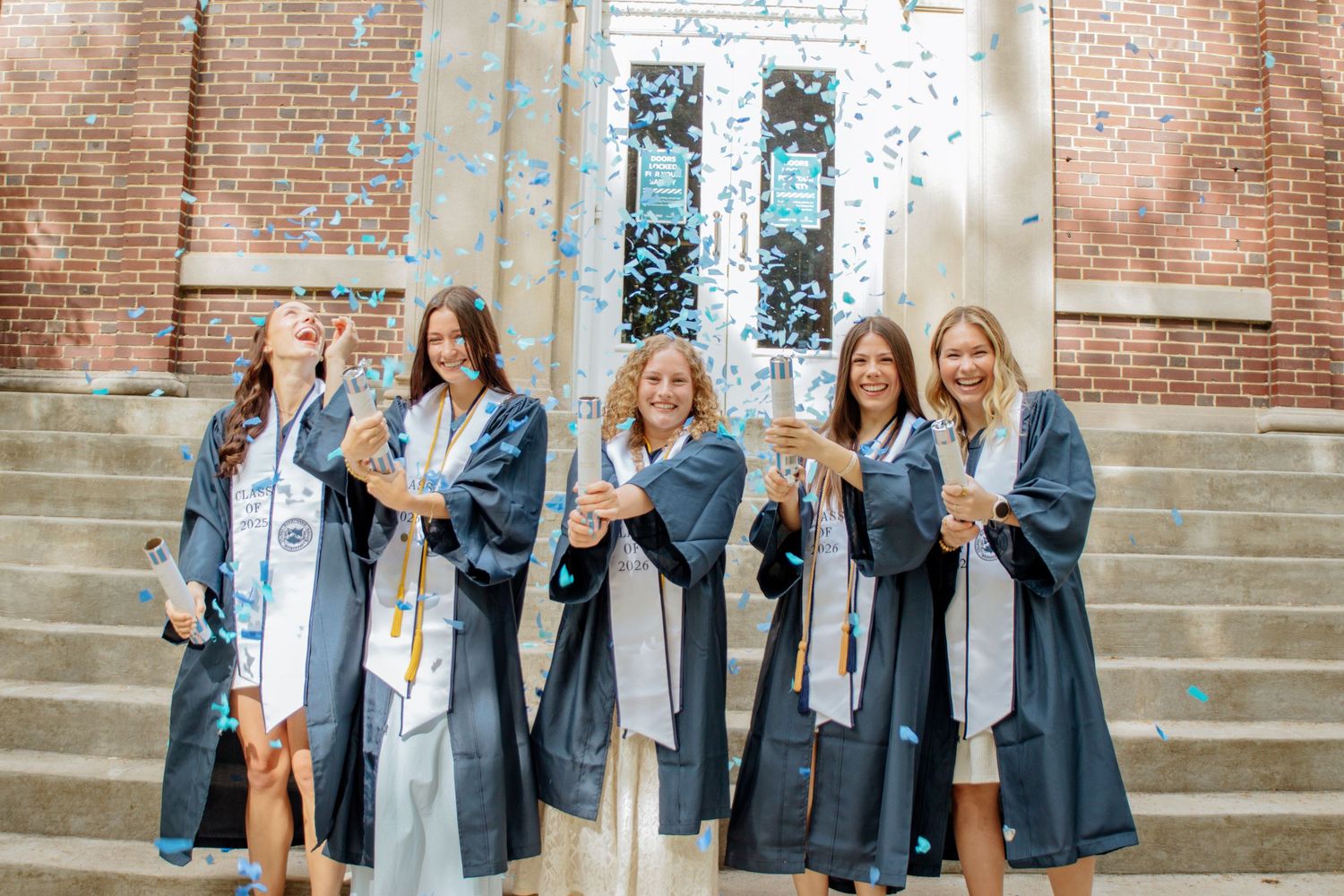 Five smiling female graduates hold champagne glasses amid falling confetti in navy graduation gowns.