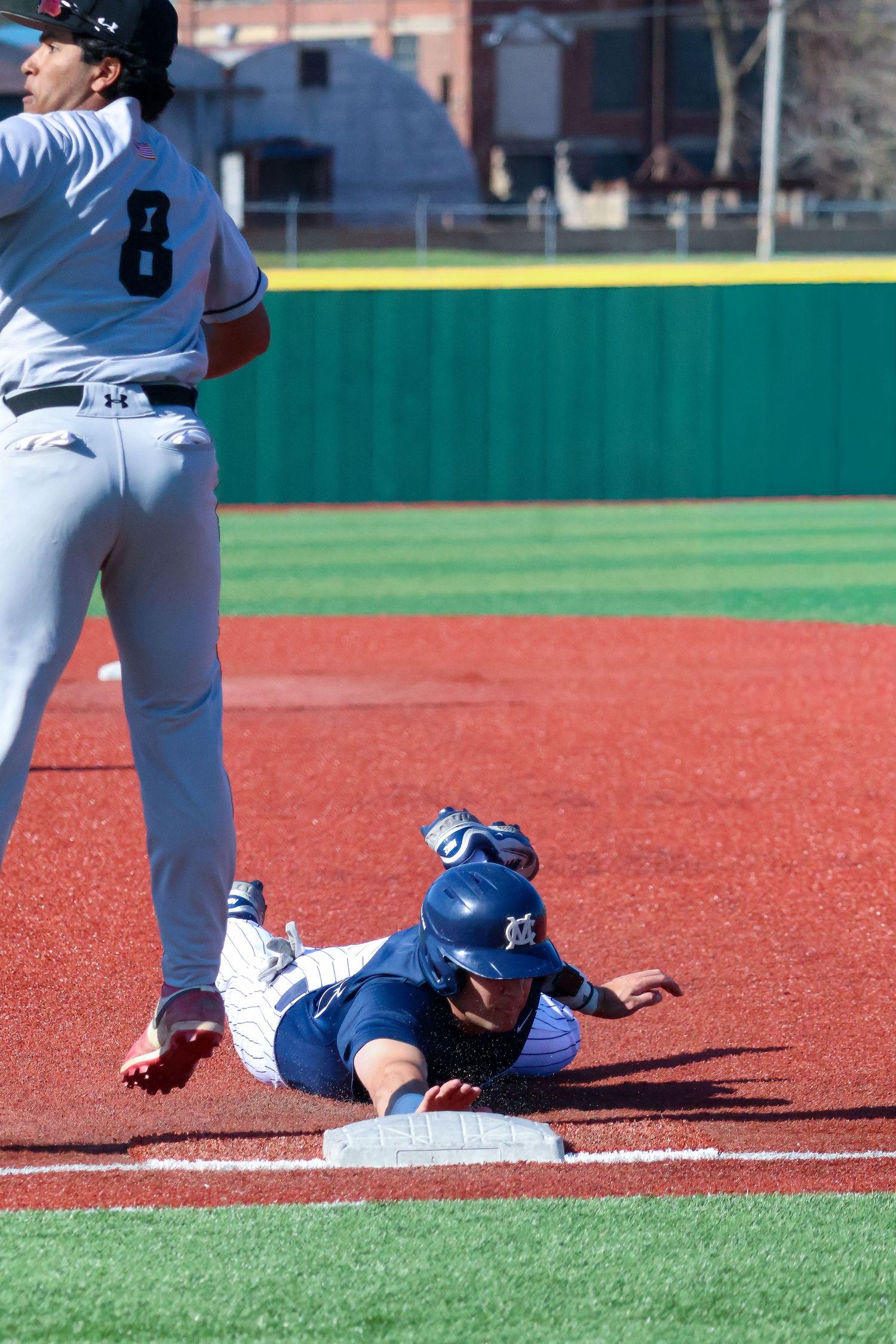 Baseball player slides headfirst into third base during competitive college game on red dirt field.