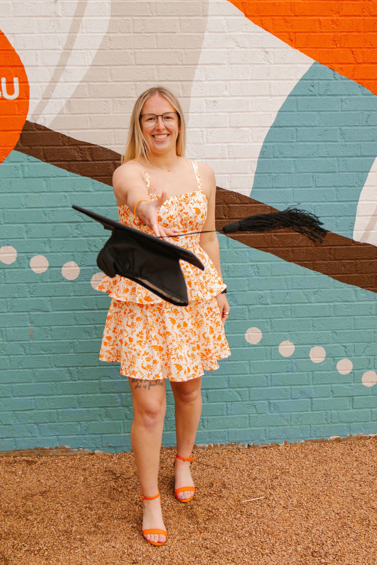 Blonde female graduate in orange floral dress holds graduation cap in front of colorful brick mural.