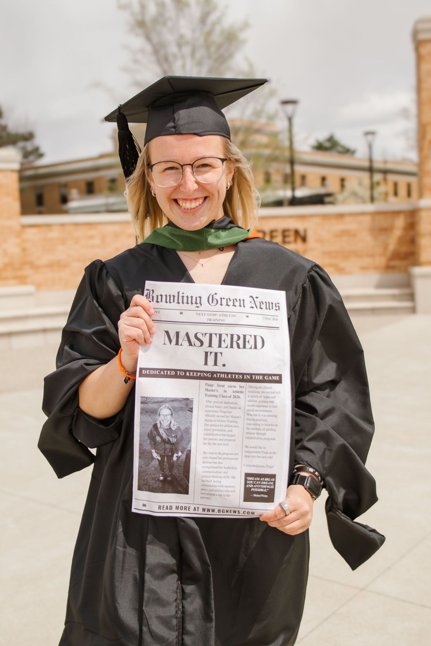 Smiling female graduate in black cap and gown holds Bowling Green News newspaper headline at BGSU.