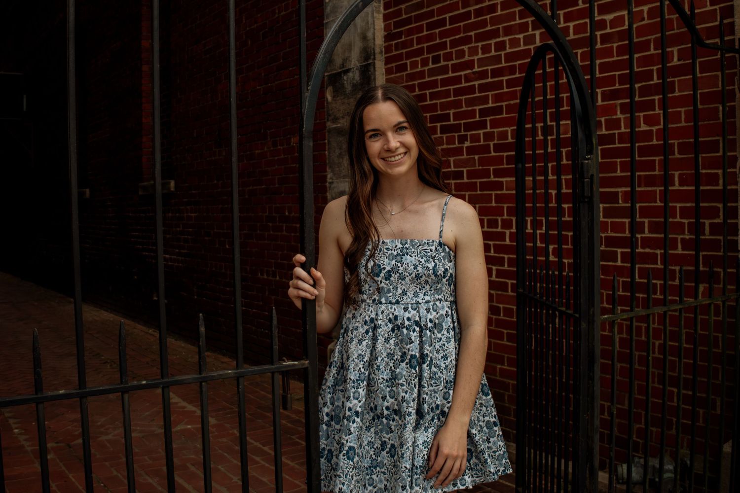 Smiling brunette woman in a blue floral dress standing by a brick wall and gate.