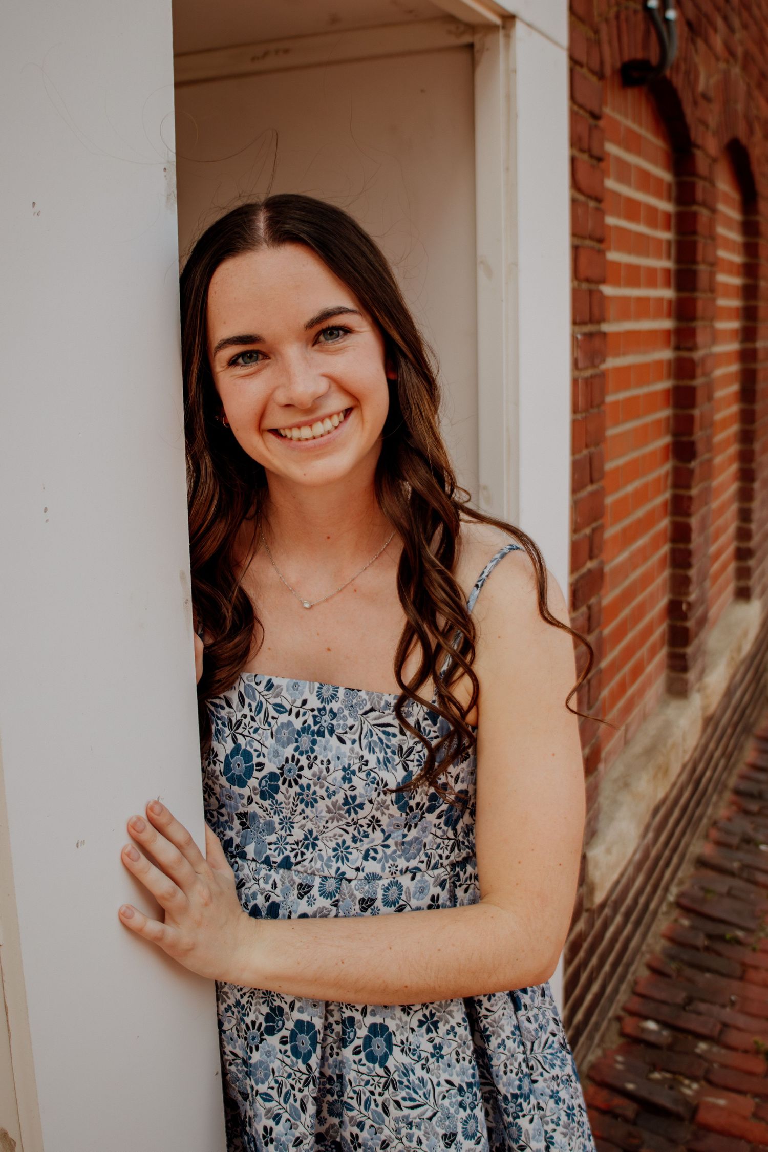 Smiling brunette woman in a blue floral strapless dress in a brick doorway.