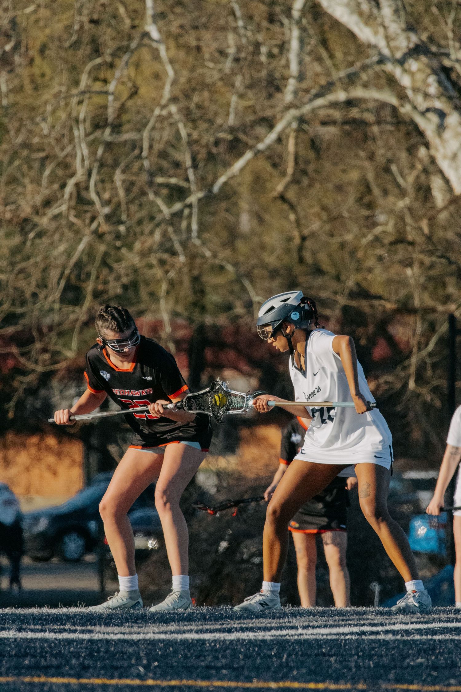 Two female lacrosse players compete for possession during an outdoor game on a sunny day.