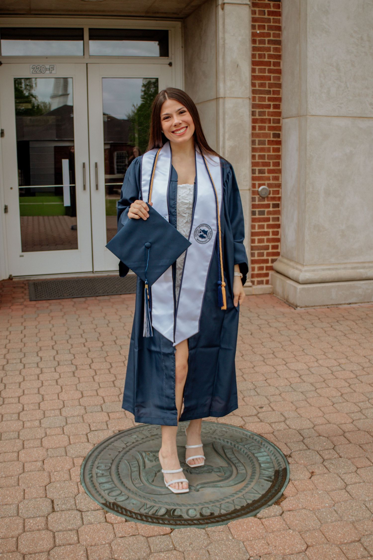 Smiling graduate in navy gown with white stole holds cap while standing on bronze university seal medallion.