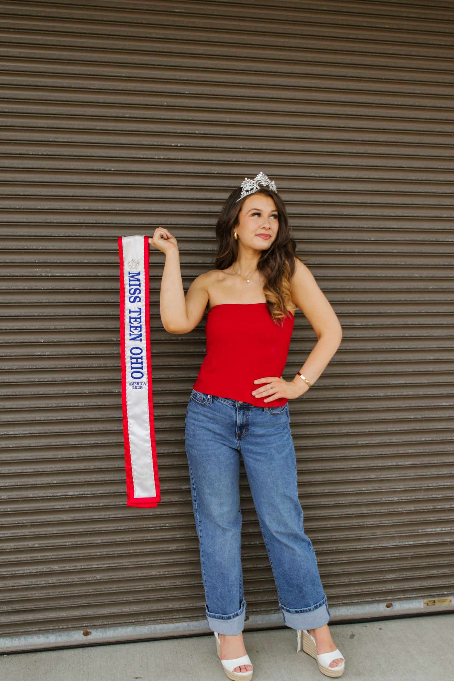 Miss Teen Ohio titleholder in red top and jeans holds sash while wearing crown outdoors.