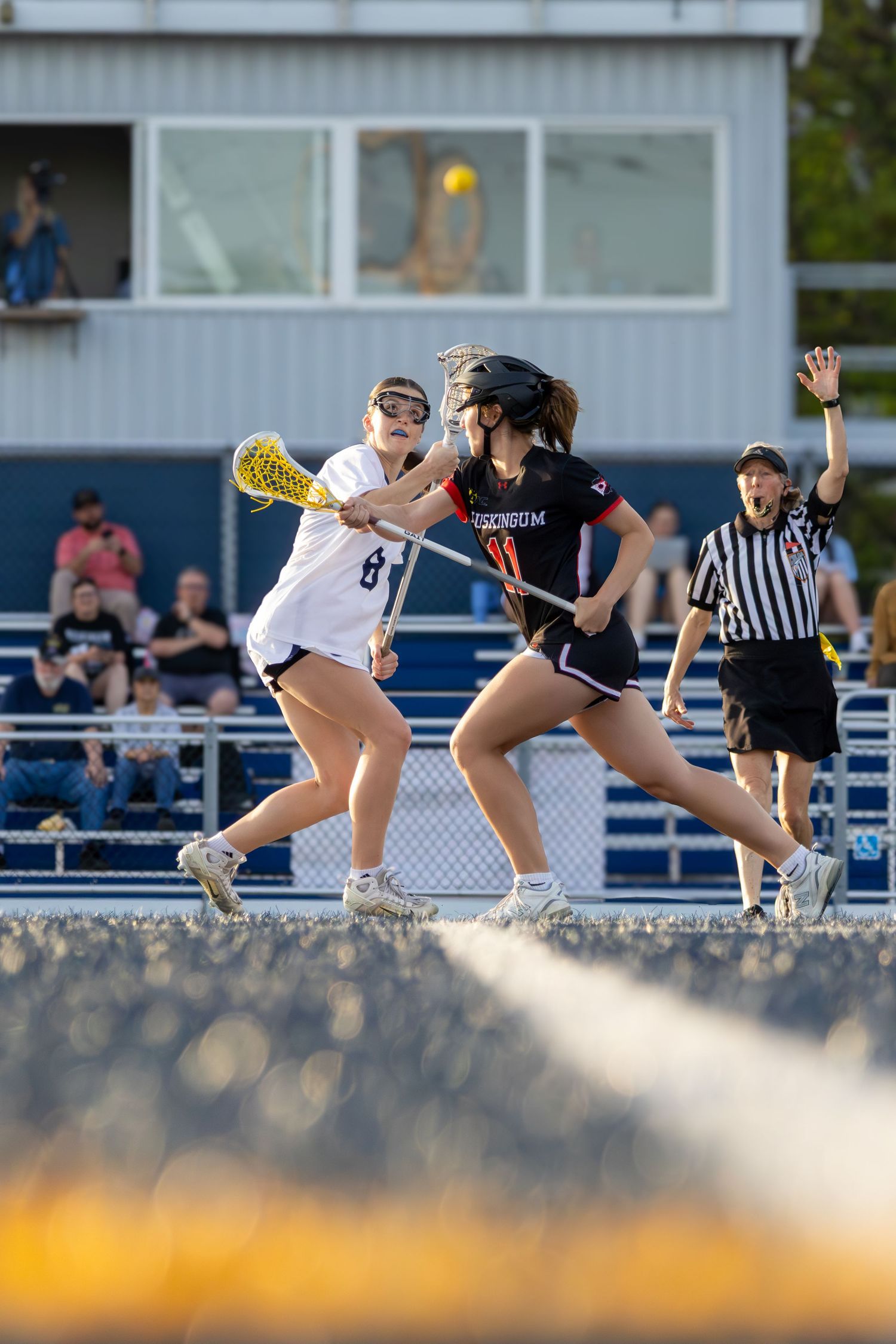 Two female lacrosse players compete for the ball as a referee signals during an outdoor high school game.