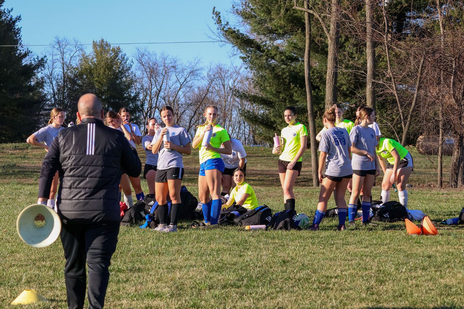 Girls soccer team listens to coach with megaphone during outdoor practice session on grass field.