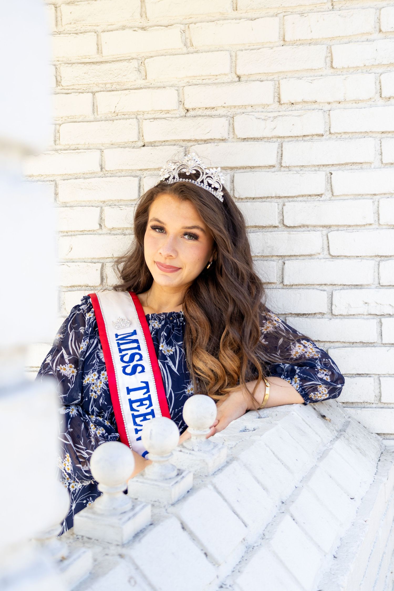 Miss Teen beauty queen in navy floral dress and crown poses pensively against white brick wall.