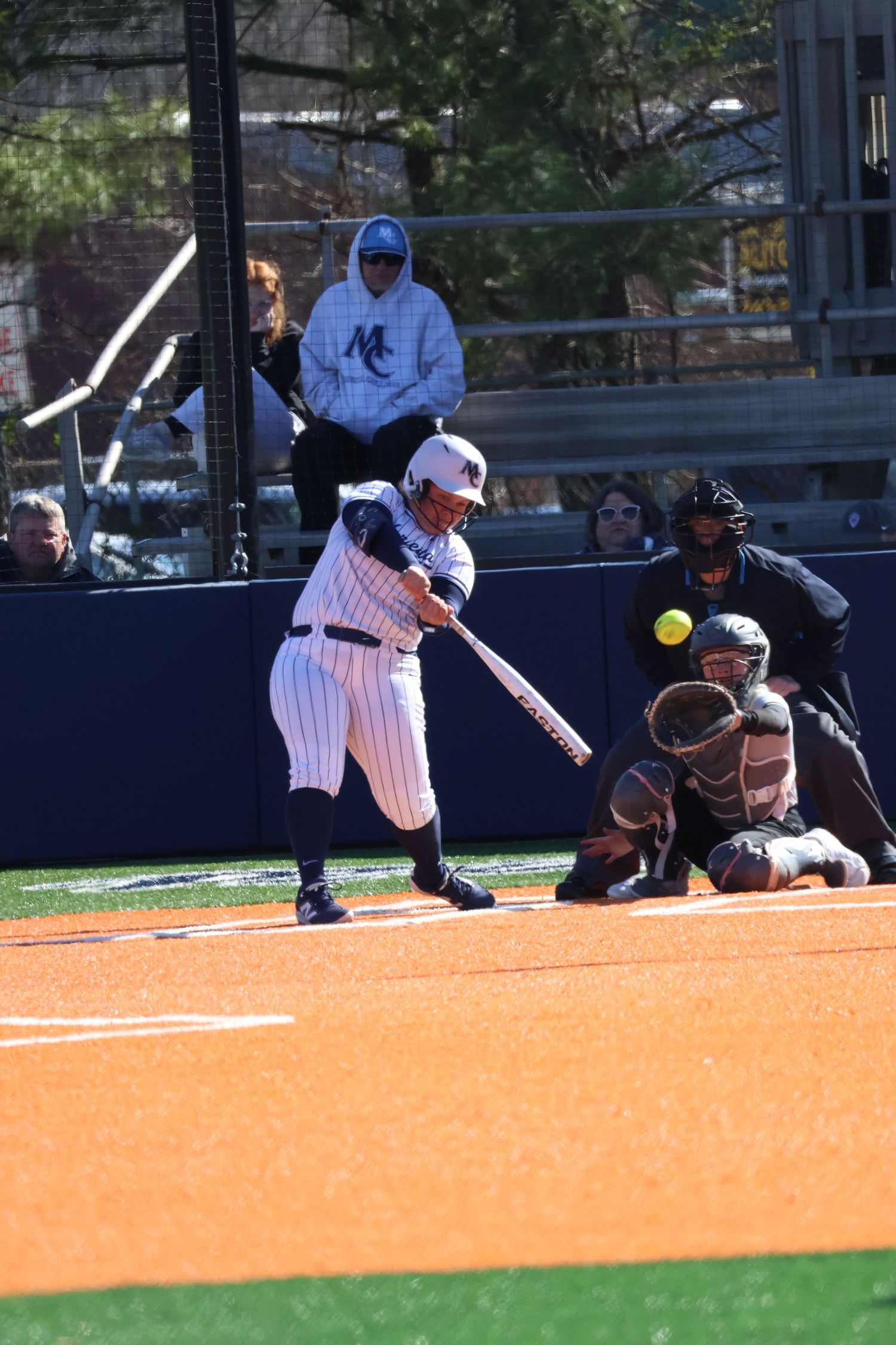 Female softball player in navy pinstripe uniform swings at a yellow ball during a college game.