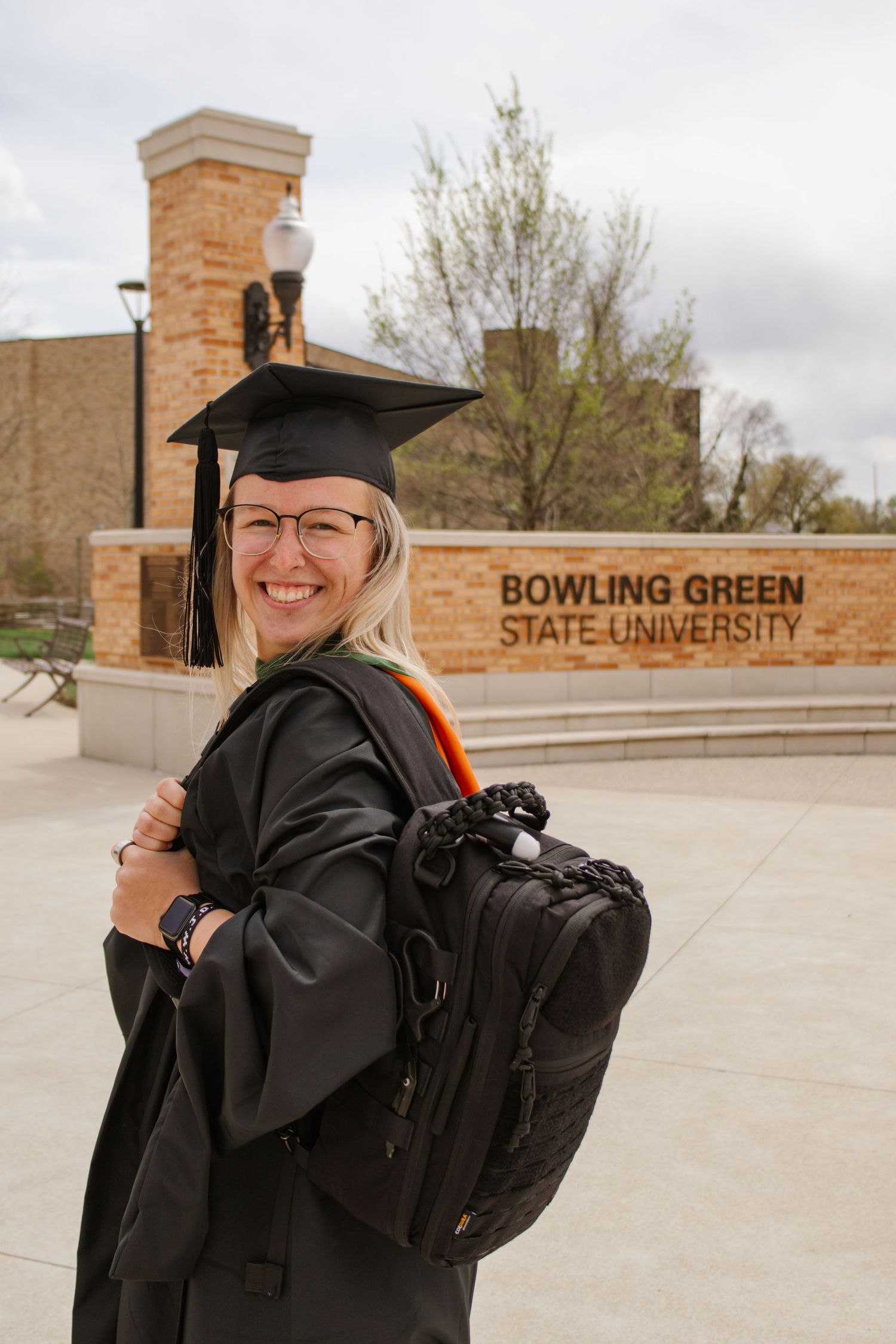 Smiling female graduate in black cap and gown with backpack poses at Bowling Green State University sign.