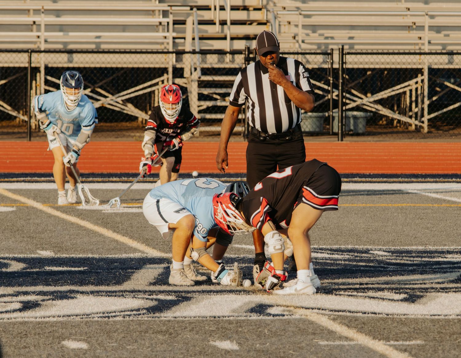 Lacrosse players compete in face-off during youth game with referee watching on stadium field.