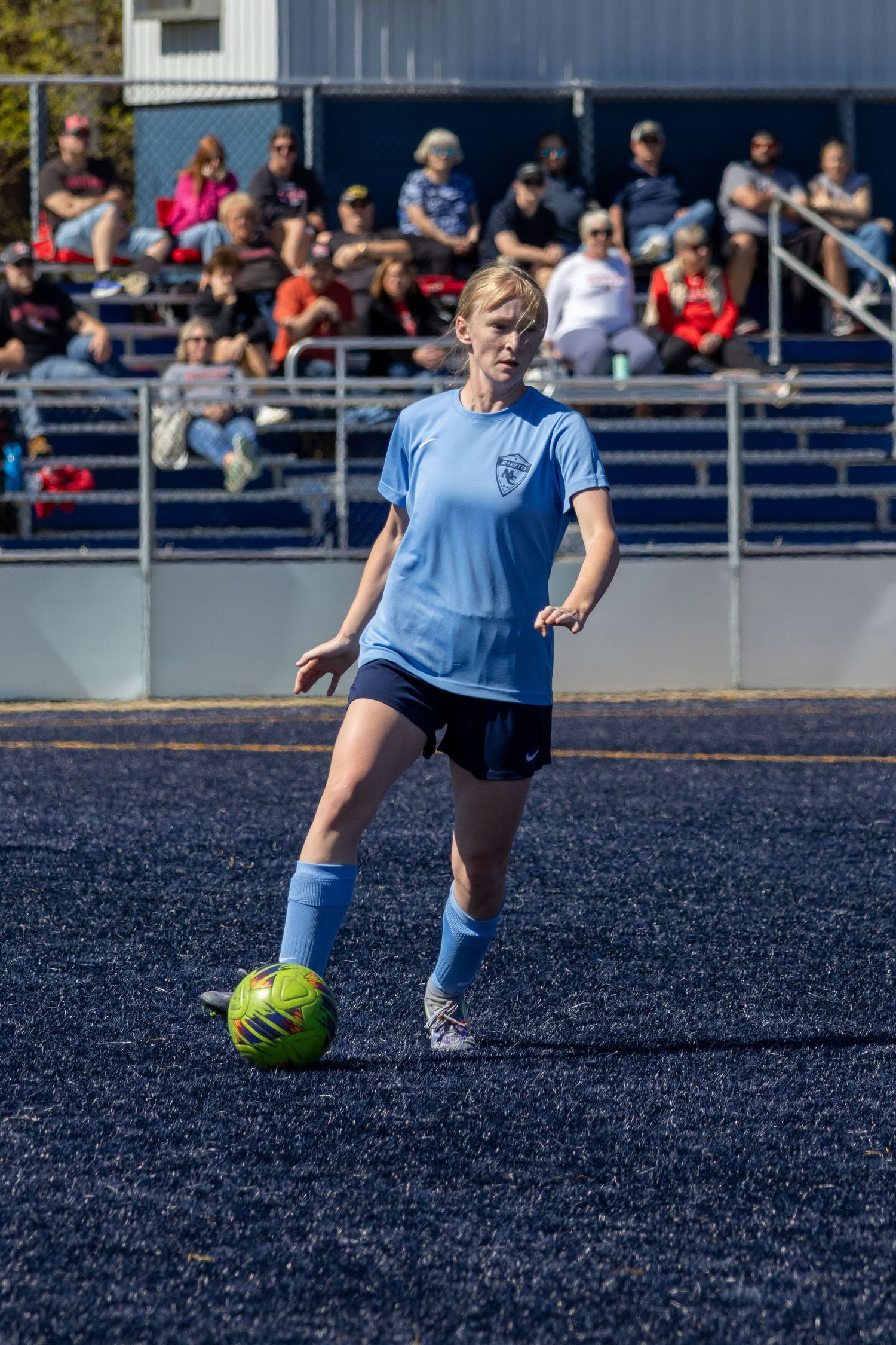 Female soccer player in light blue uniform dribbles neon green ball on dark turf field.