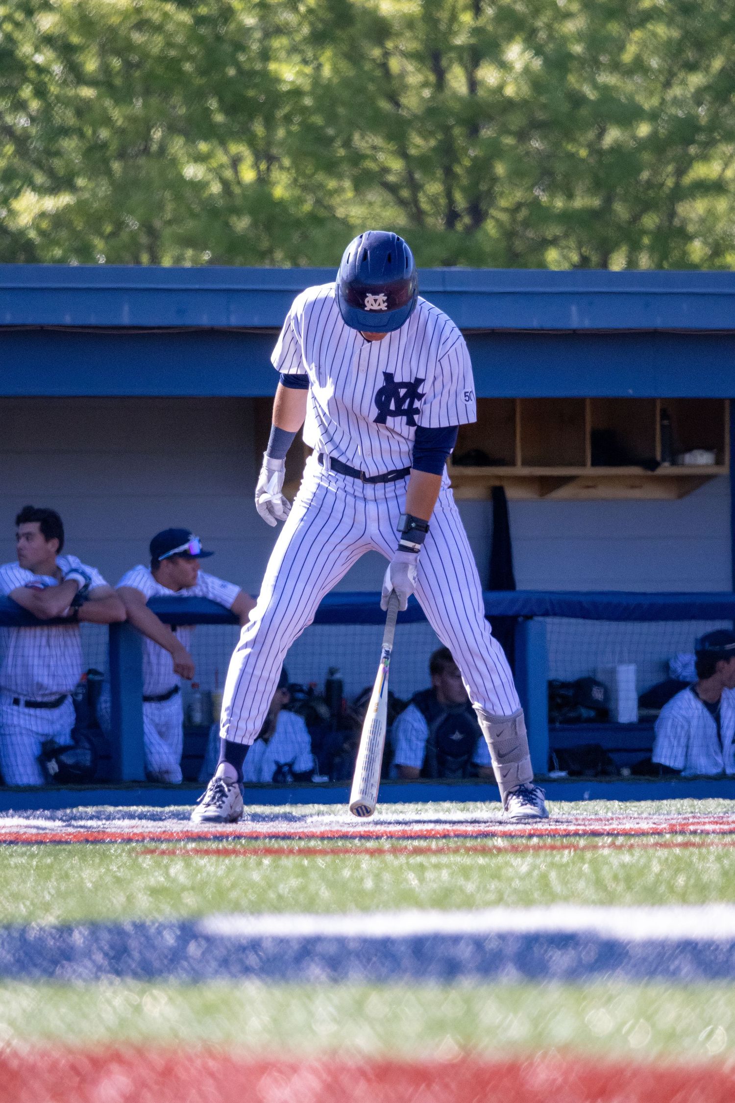 Baseball player in pinstripe uniform stands at bat, ready to swing, with dugout and teammates visible in background.
