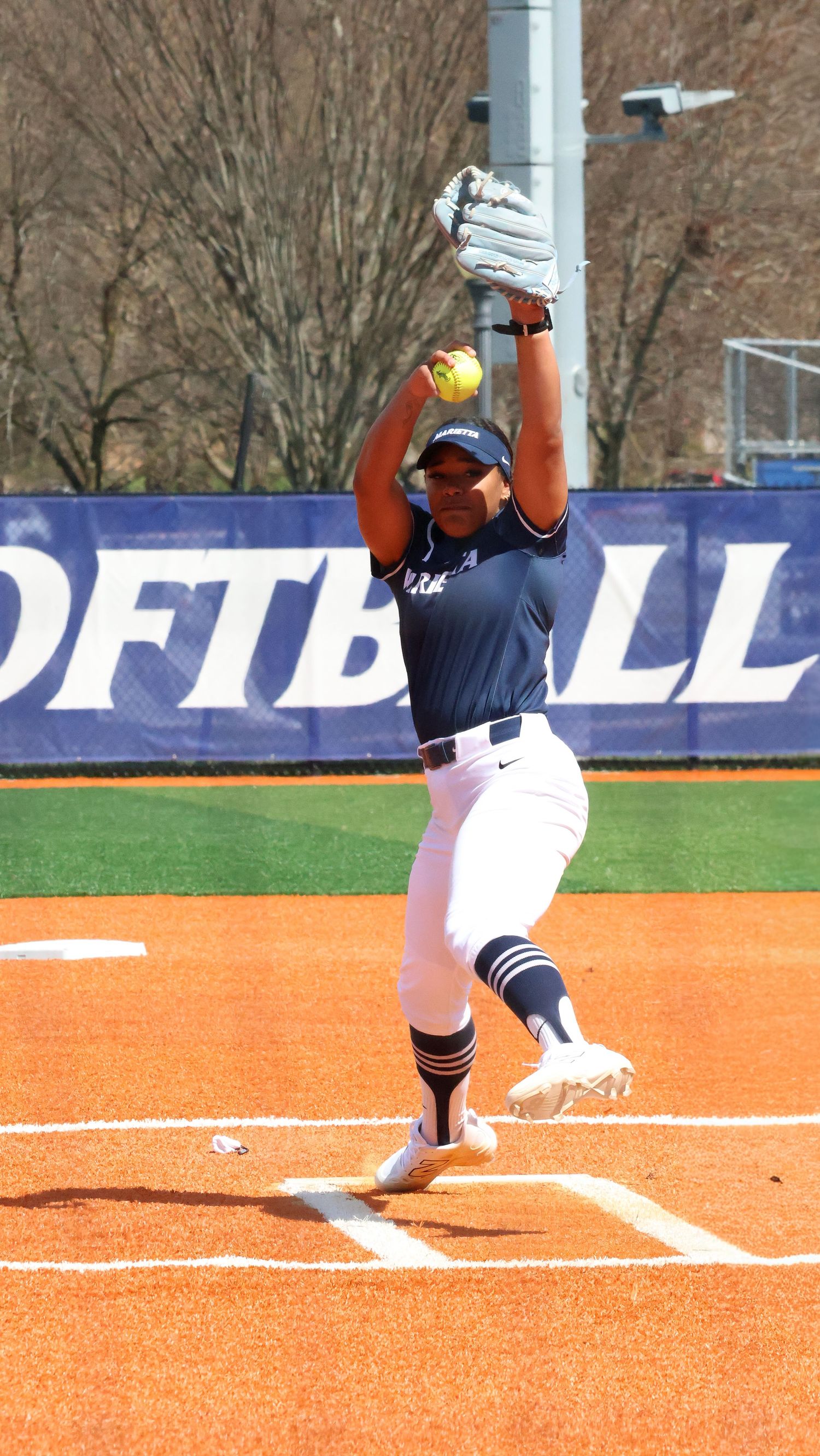 Female softball pitcher in navy uniform mid-windup on red clay field with SOFTBALL banner in background.